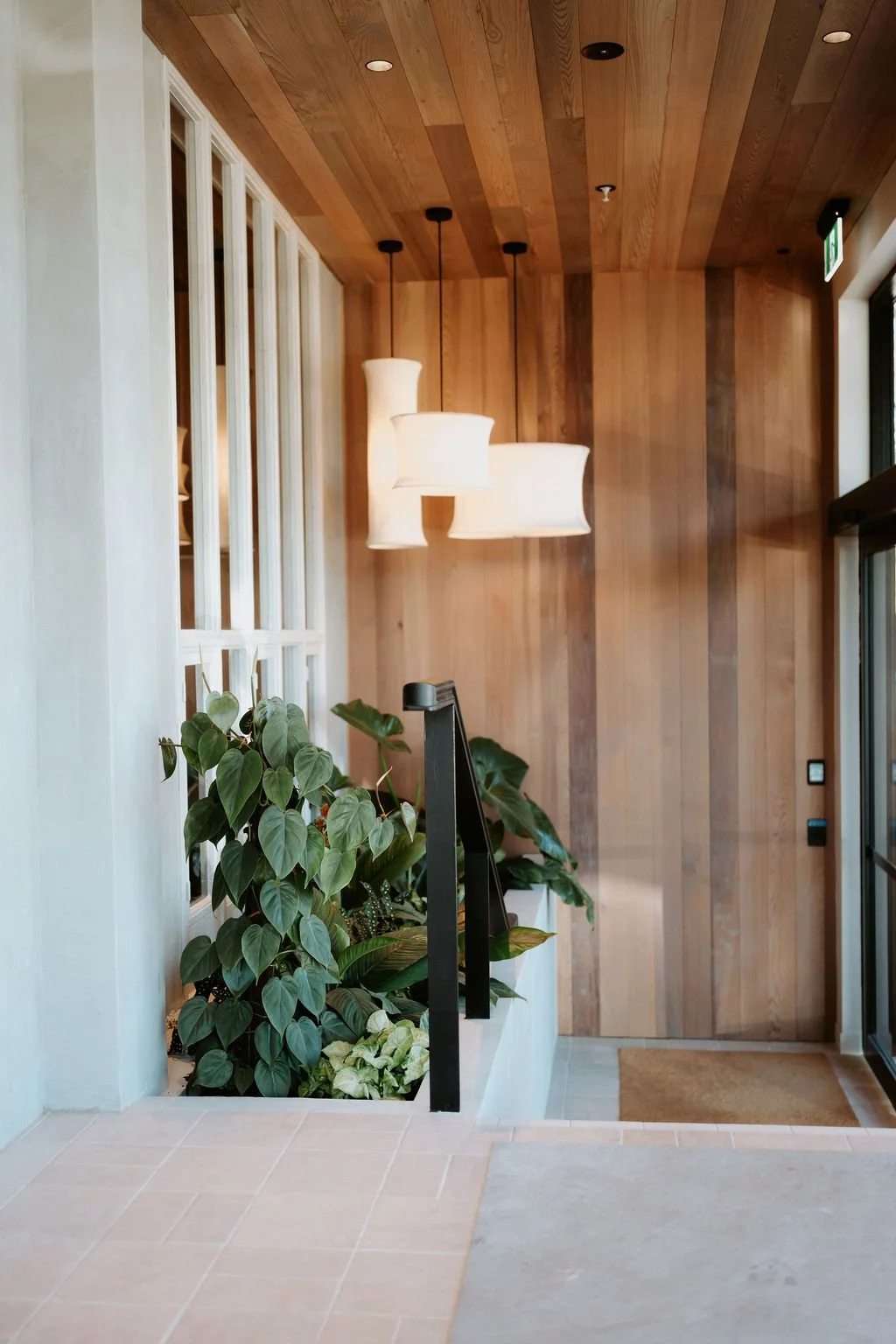 Interior of a modern space with wooden walls and ceiling, hanging white pendant lights, indoor plants, a glass door, and a floor mat.
