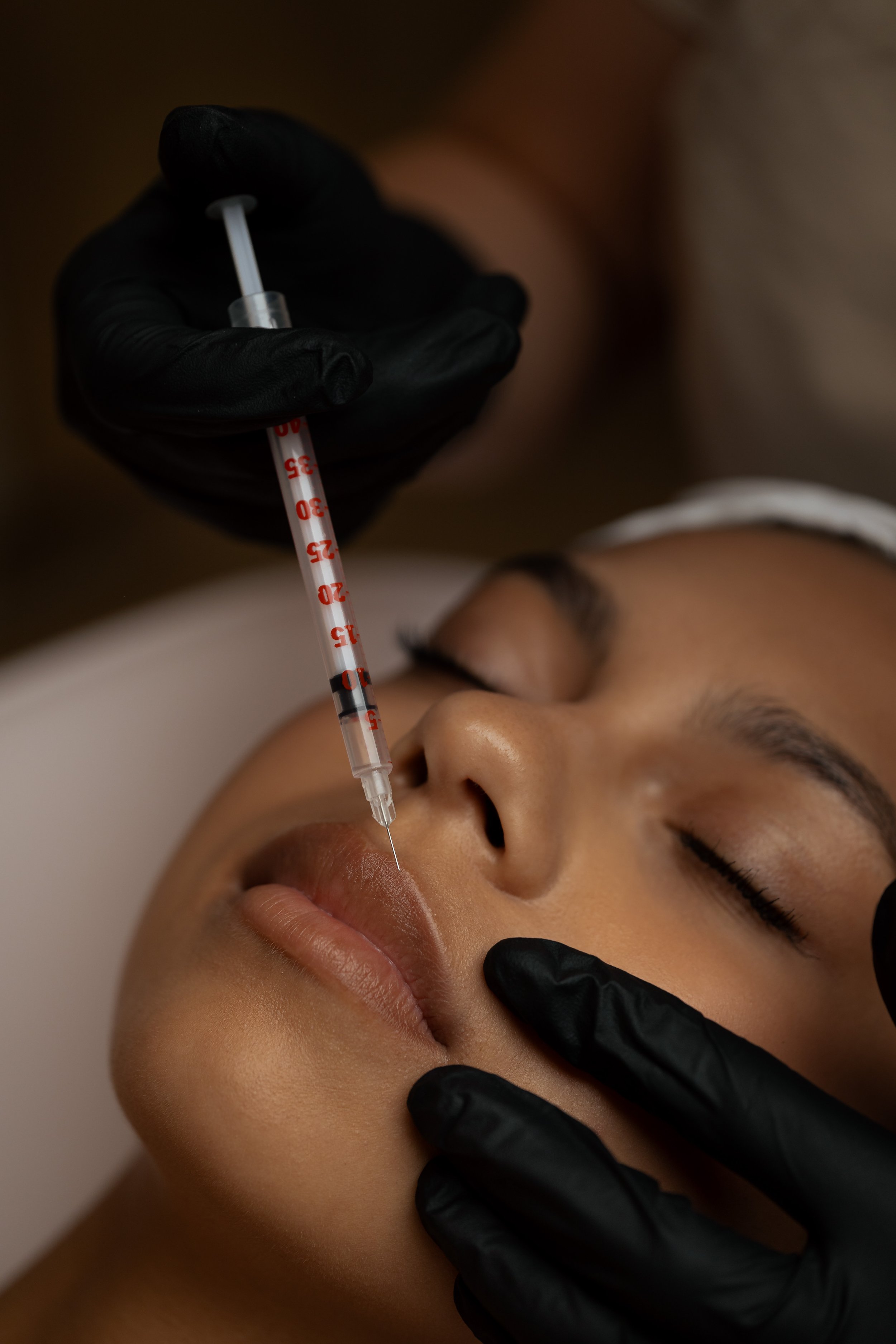 A person receiving a facial treatment with a syringe held by a practitioner wearing black gloves. The person has closed eyes with makeup visible on the eyelids.