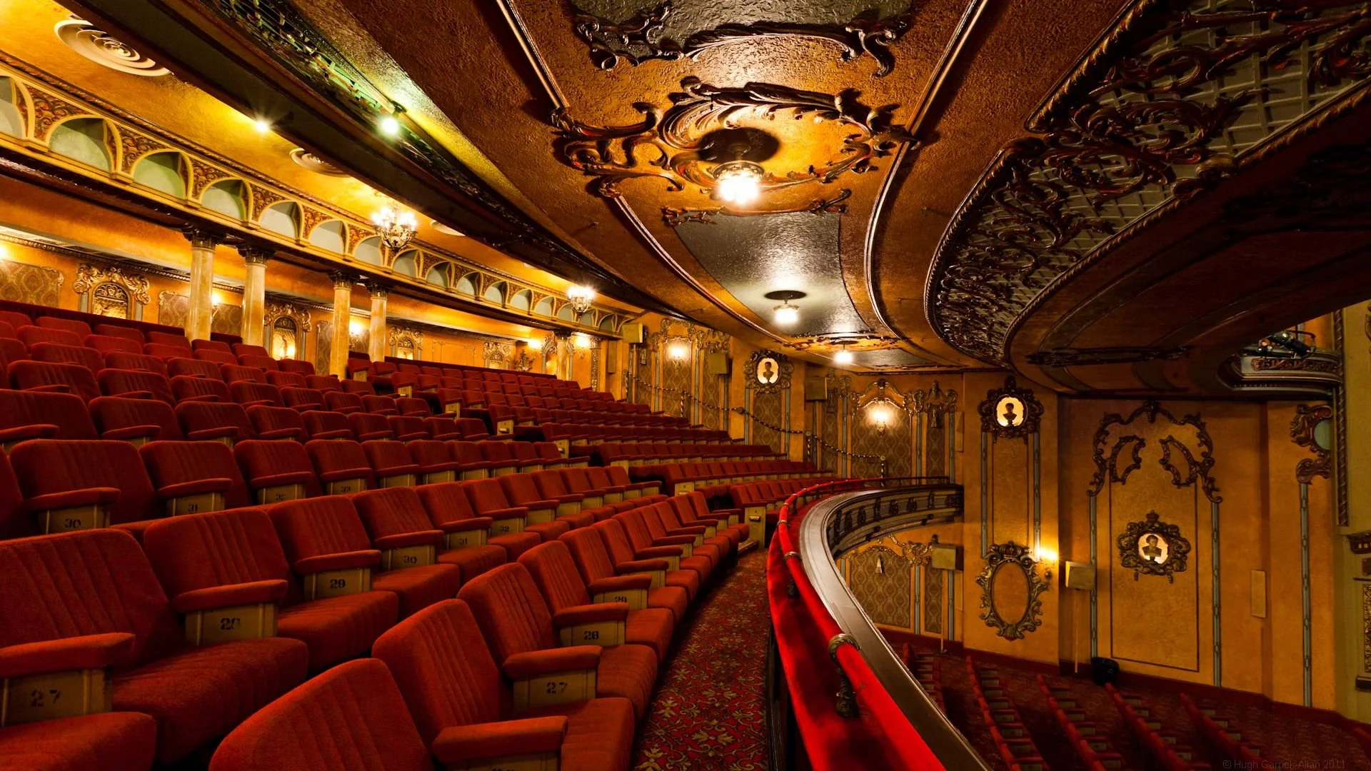 An ornate theater with red velvet seats, gold and dark wood decorative walls, and a curved balcony with intricate black iron railings.