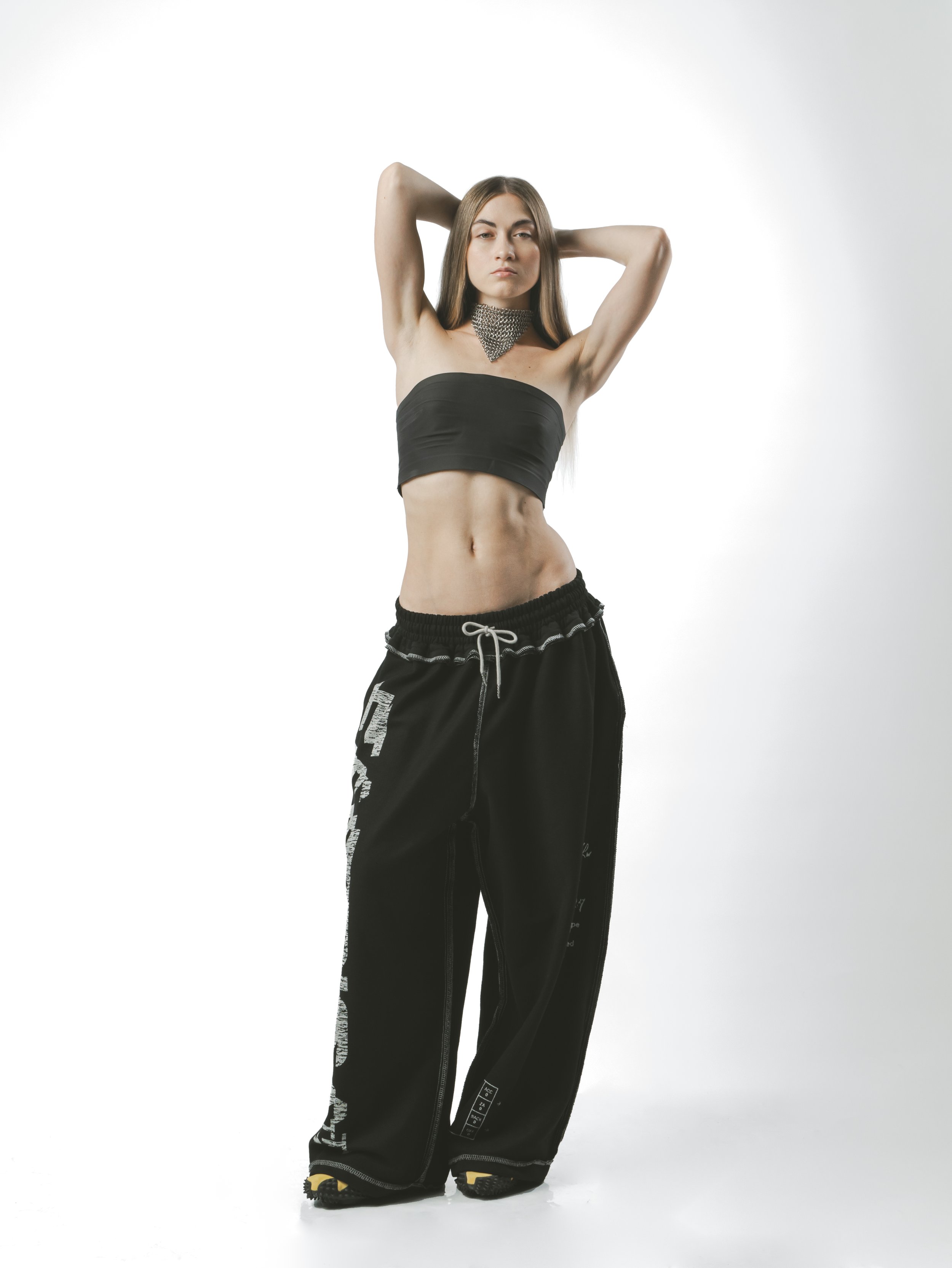 Young woman with long hair wearing a black tube top, baggy black pants, and yellow shoes stands against a white background, with her arms raised and hands behind her head, looking confidently at the camera.