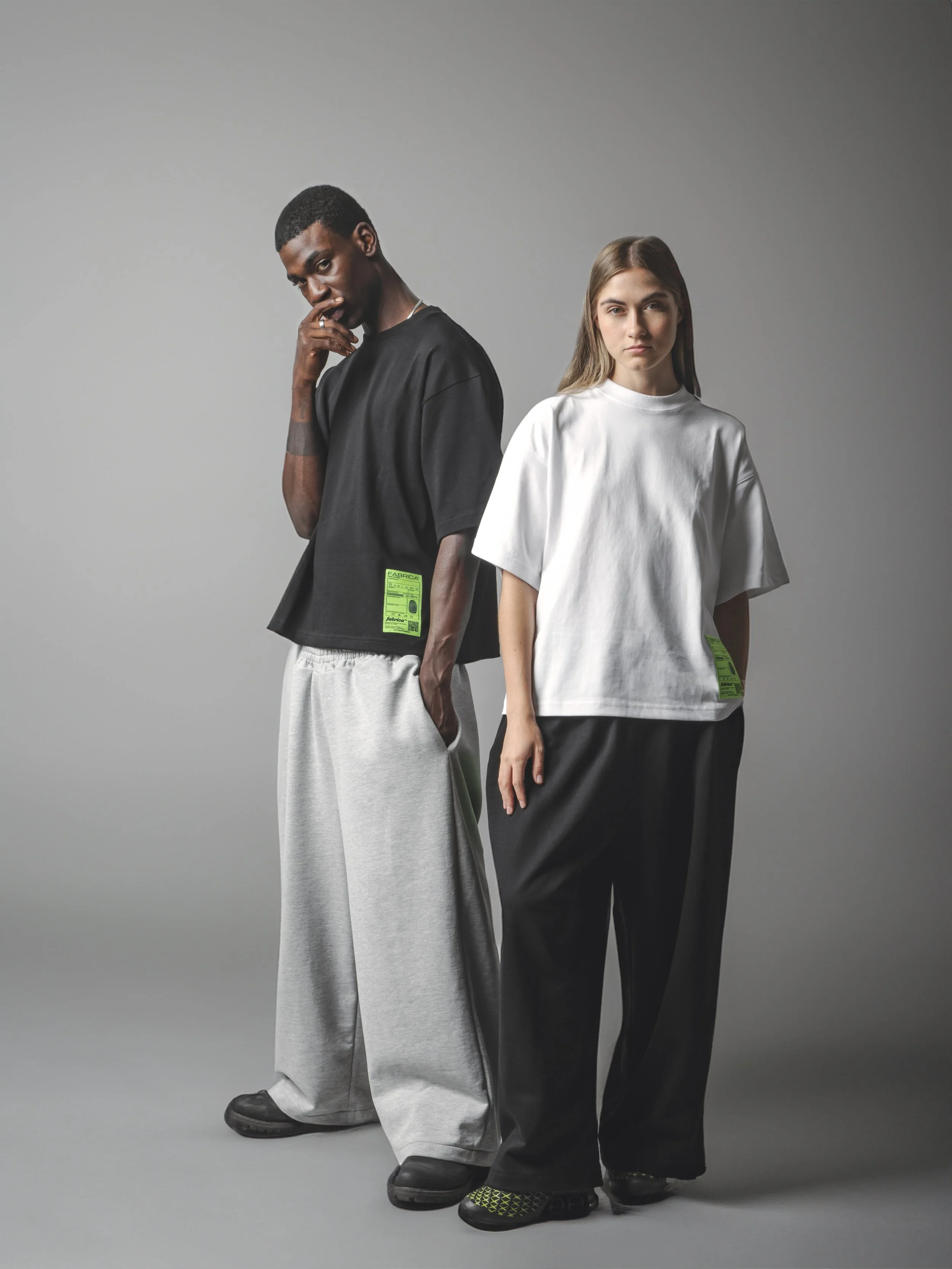 Two young adults standing side by side against a plain grey background. The person on the left is a Black male with short hair, wearing a black oversized t-shirt with a green label on the sleeve, light-colored wide-leg pants, and black shoes. The per