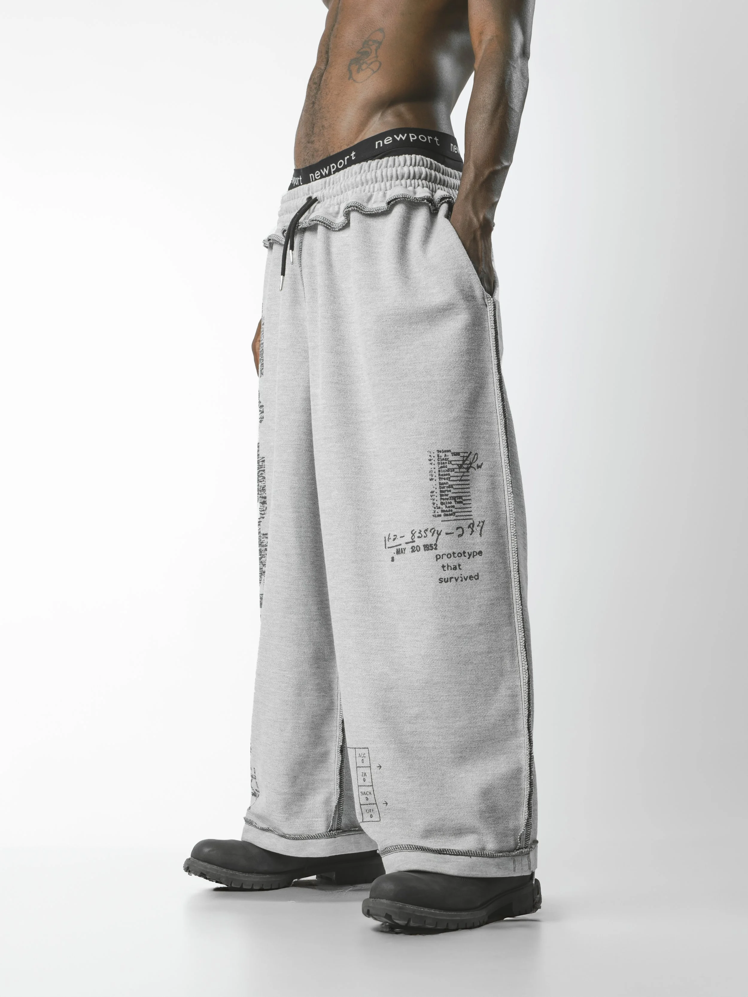 Close-up of a person's lower body wearing light gray sweatpants with printing on the side and black boots, standing against a plain white background.