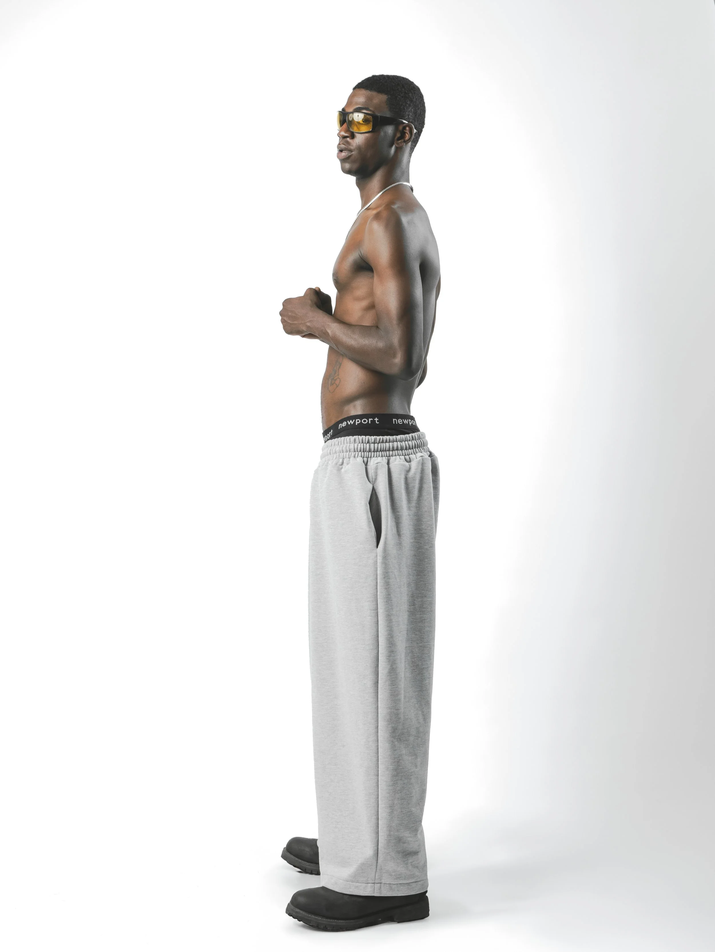 A man standing in profile, shirtless, wearing gray sweatpants with a black waistband labeled 'newport,' black boots, yellow-tinted sunglasses, and a silver chain necklace against a white background.