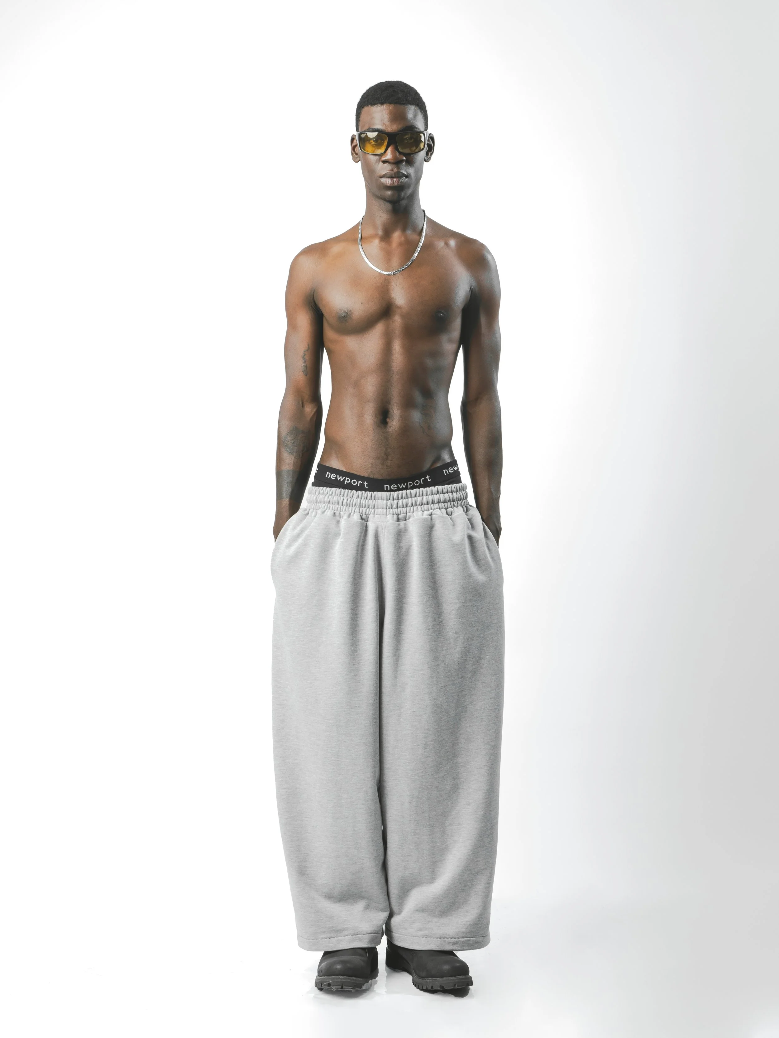A shirtless man in sunglasses, wearing gray sweatpants, black boots, a silver chain necklace, standing against a white background.
