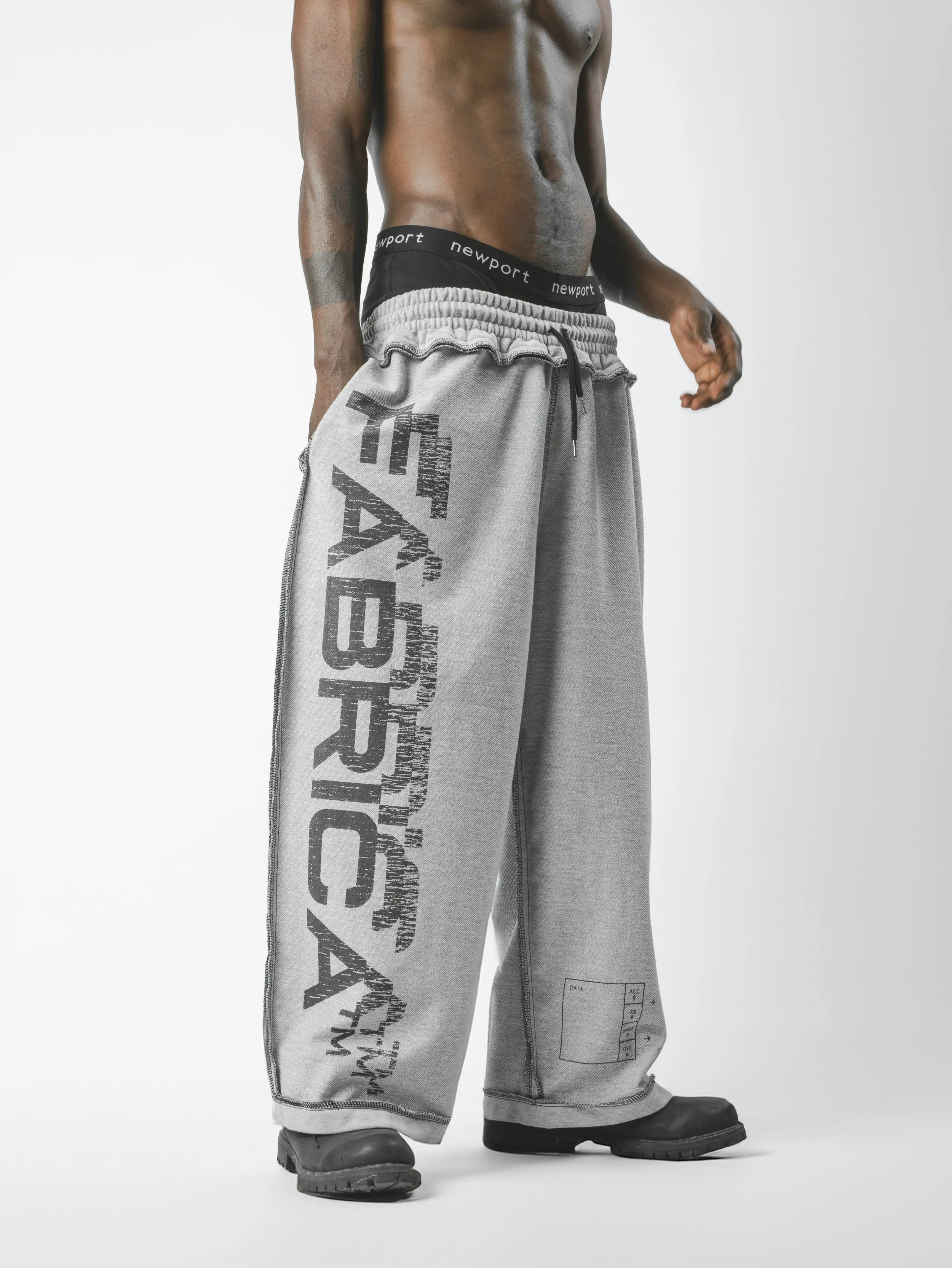 Close-up of a male figure with tattoos wearing gray sweatpants with black text and logo, black boots, and a white background.