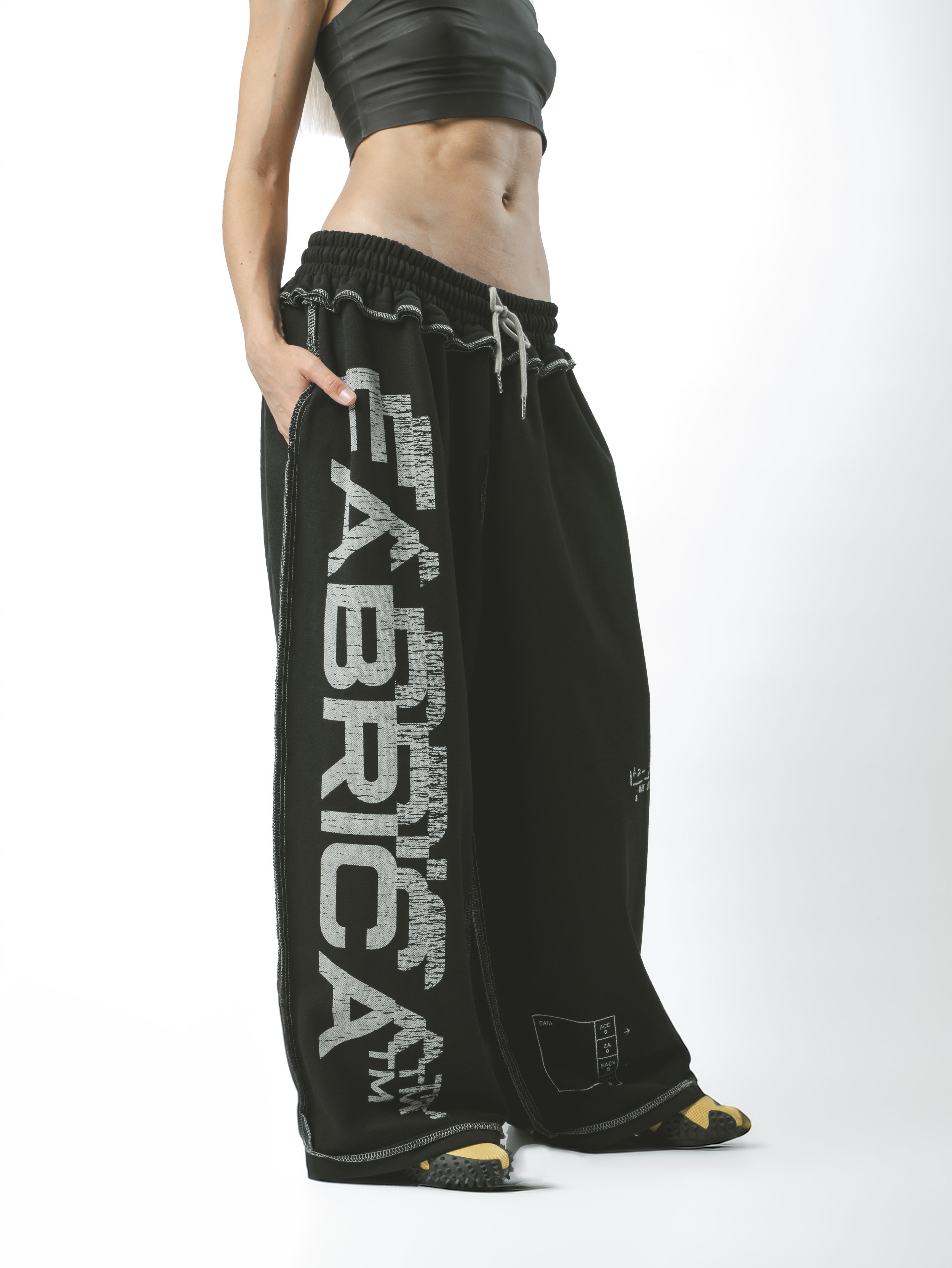 Person wearing black graphic sweatpants and yellow and black sneakers, with a cropped athletic top, standing against a white background.