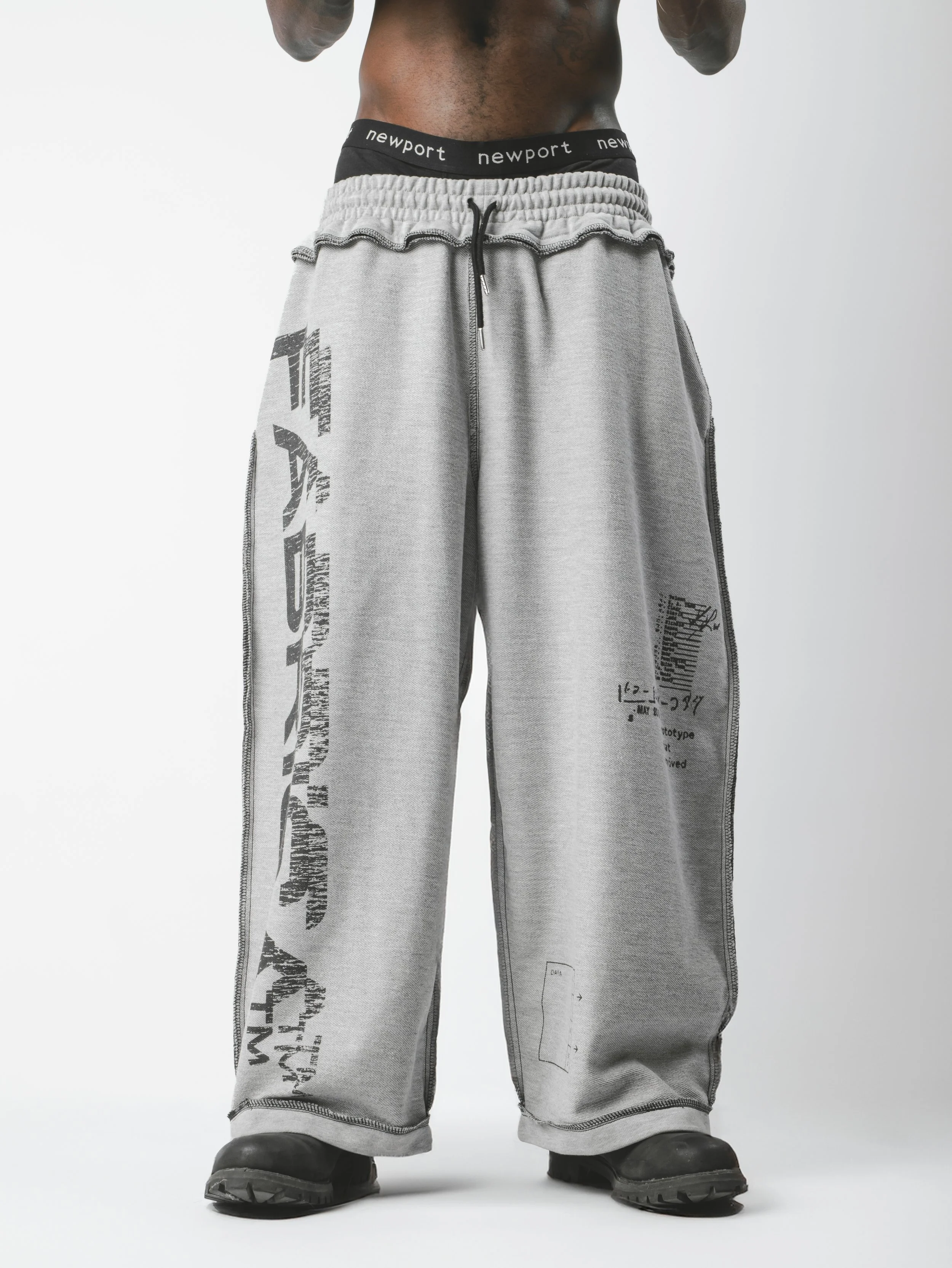 Close-up of the lower torso and legs of a person wearing gray sweatpants with black text and graphics, black shoes, and black underwear with white text.