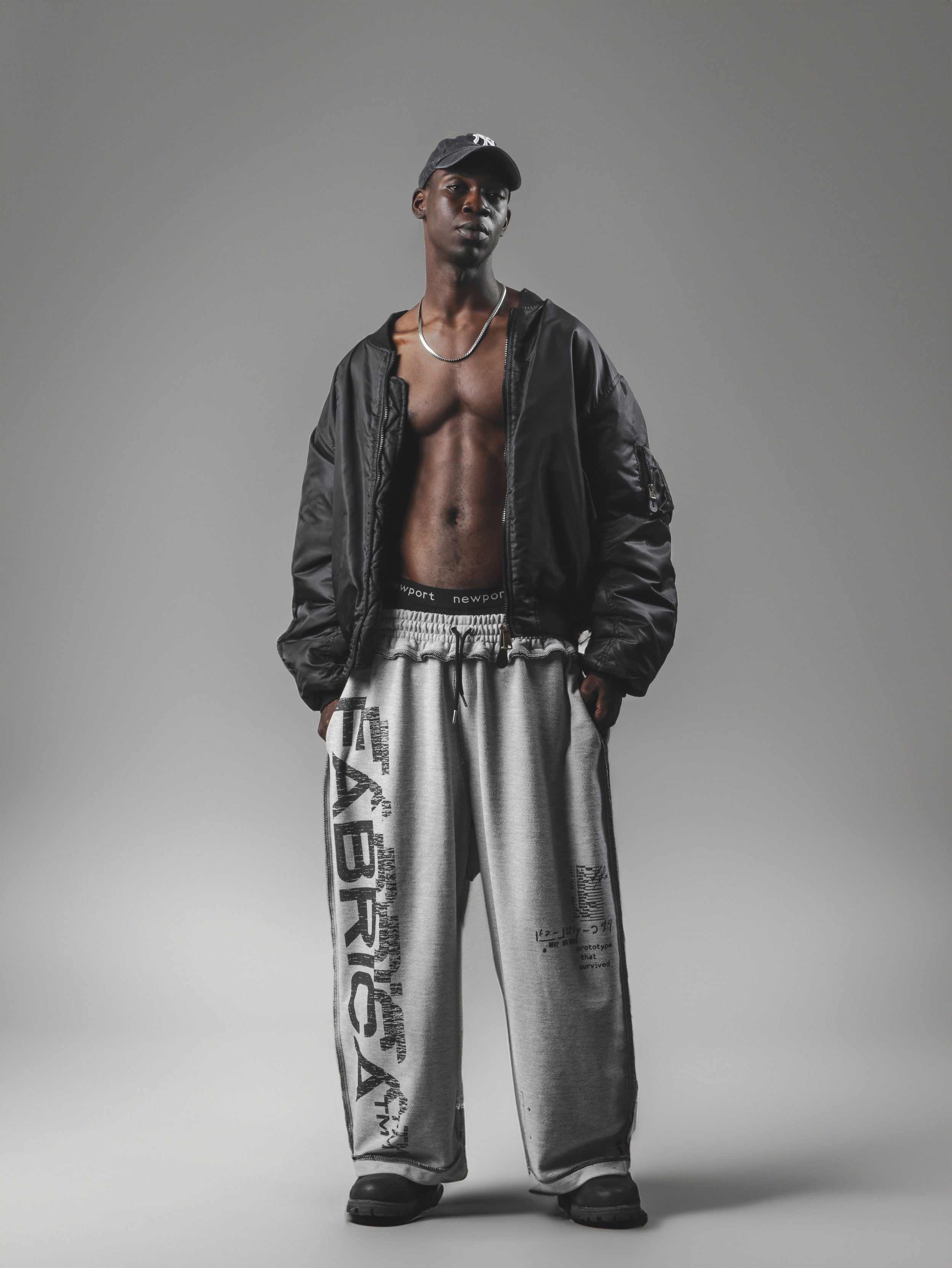 A young man with dark skin and an athletic build standing against a plain gray background, wearing a black baseball cap, a black jacket with the front open revealing his bare chest, baggy gray sweatpants featuring black and white graphic print on the