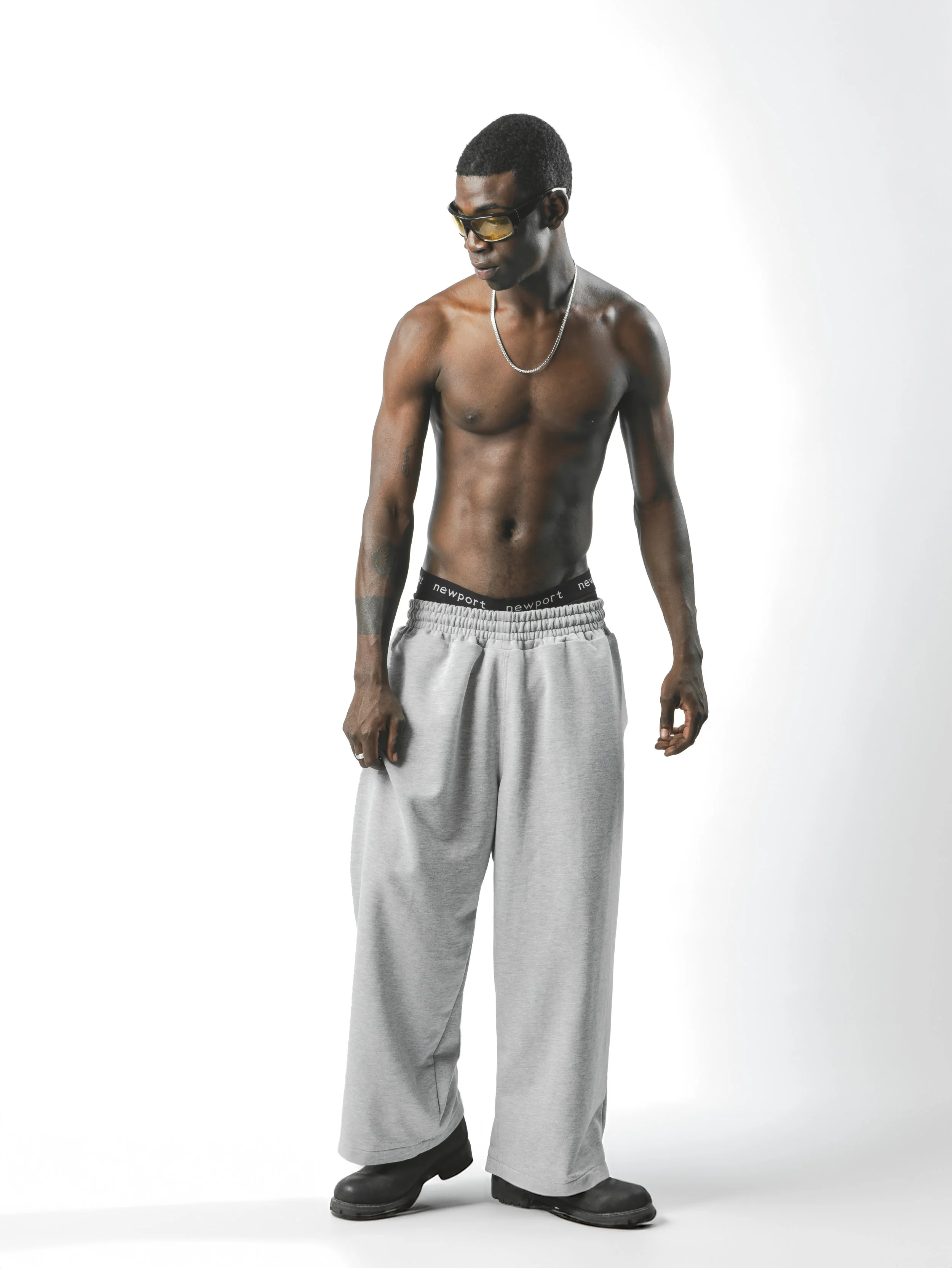 A featureless mannequin with dark skin, short hair, dark sunglasses, and a silver chain necklace, wearing gray sweatpants and black boots, stands against a plain white background.
