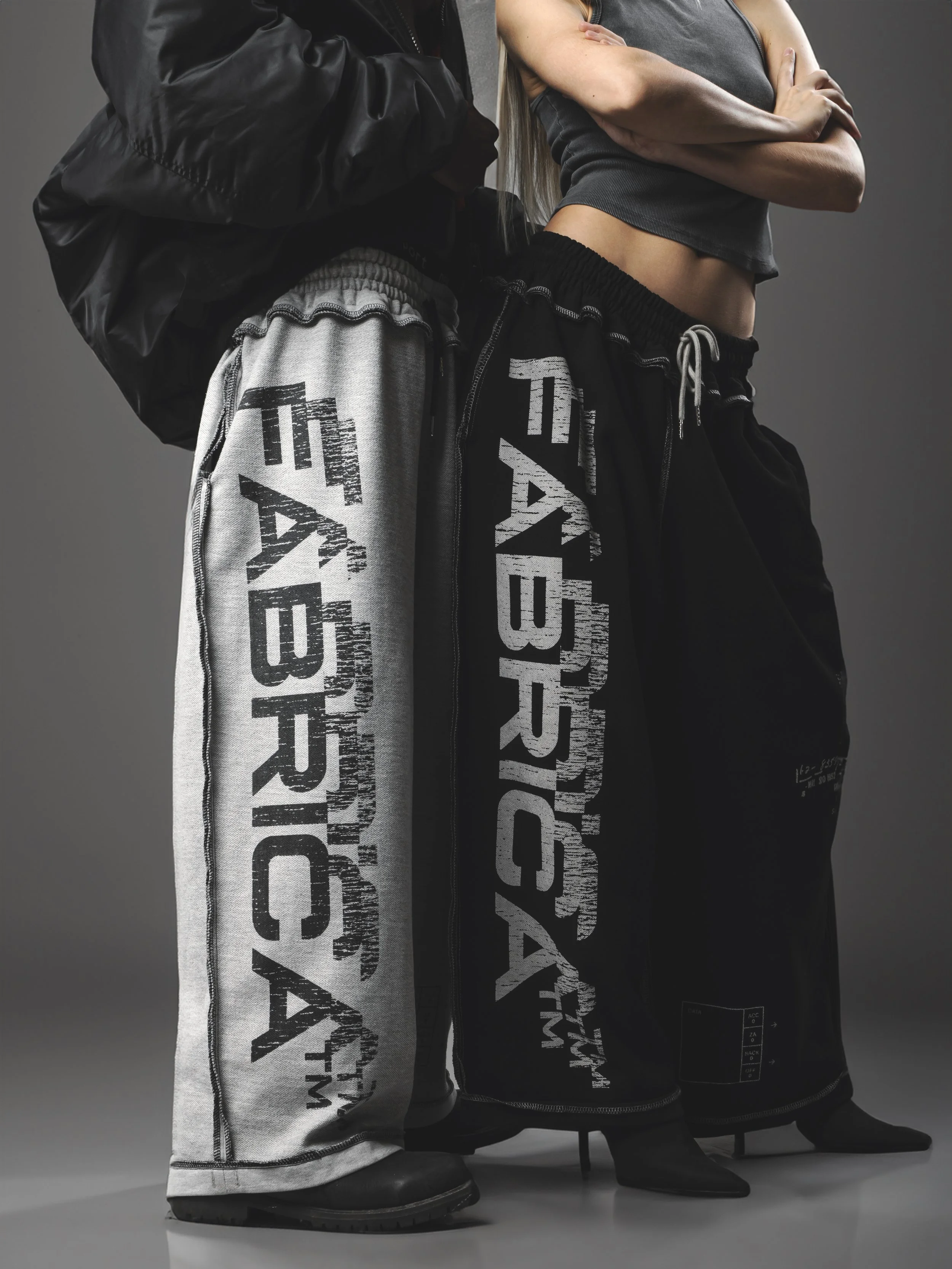 Close-up of three people standing in a row, showing their legs and footwear. They are wearing different pants with bold, distressed text and graphics, along with shoes like boots and pointed-toe heels, against a plain gray background.