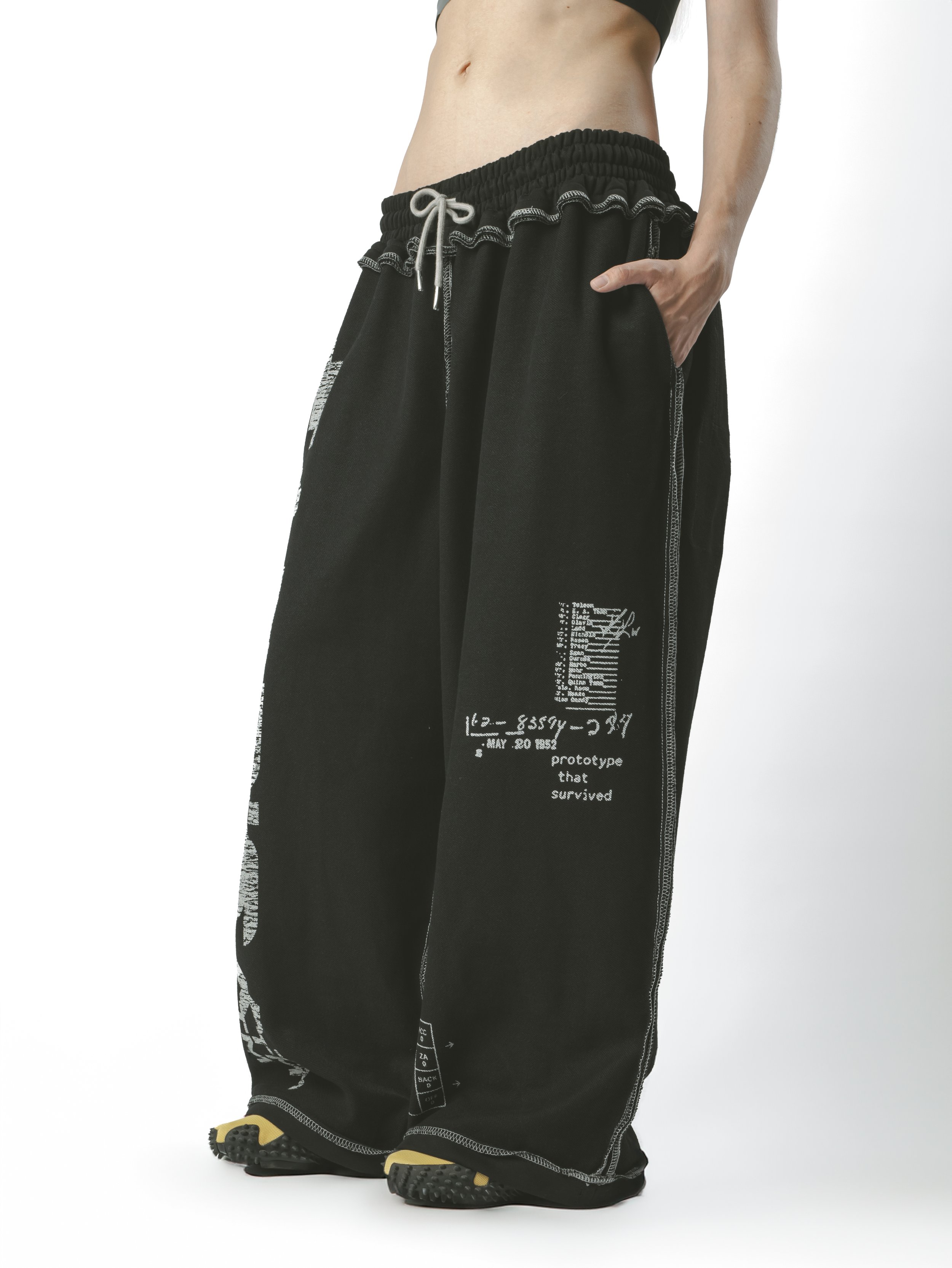 Person wearing black oversized pants with white text and symbols, black shoes with yellow soles, and a black top, standing against a plain white background.