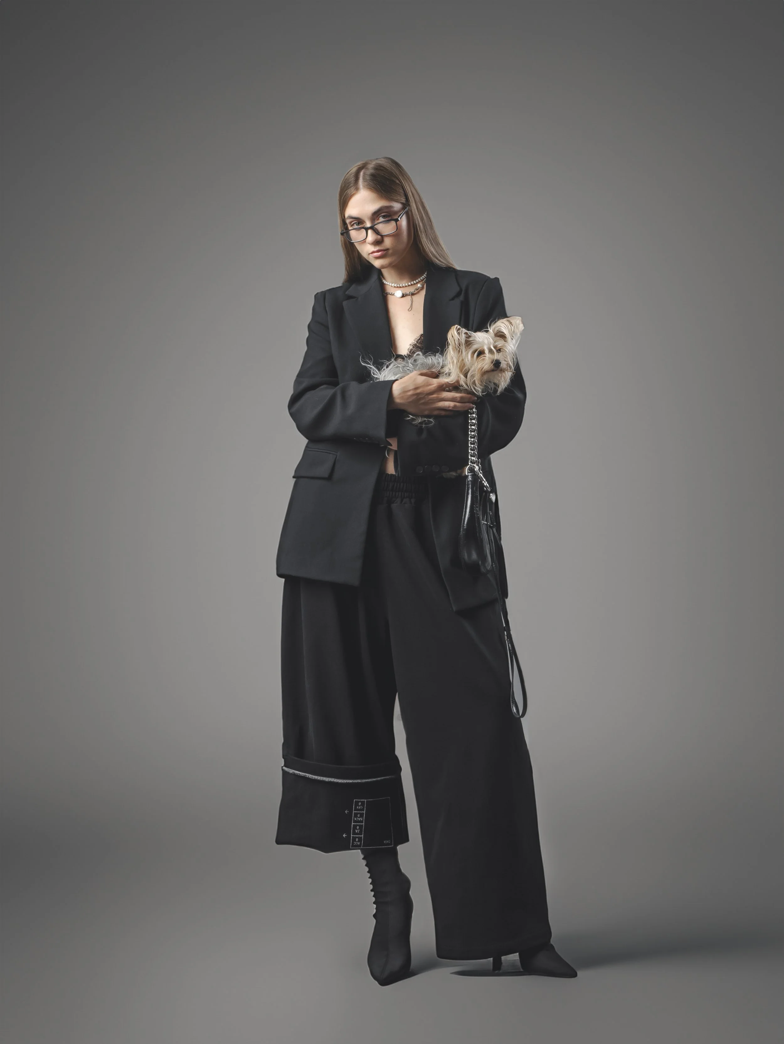 A young woman with long brown hair and glasses wearing a black blazer and black pants, holding a small Yorkshire Terrier dog, standing against a plain gray background.