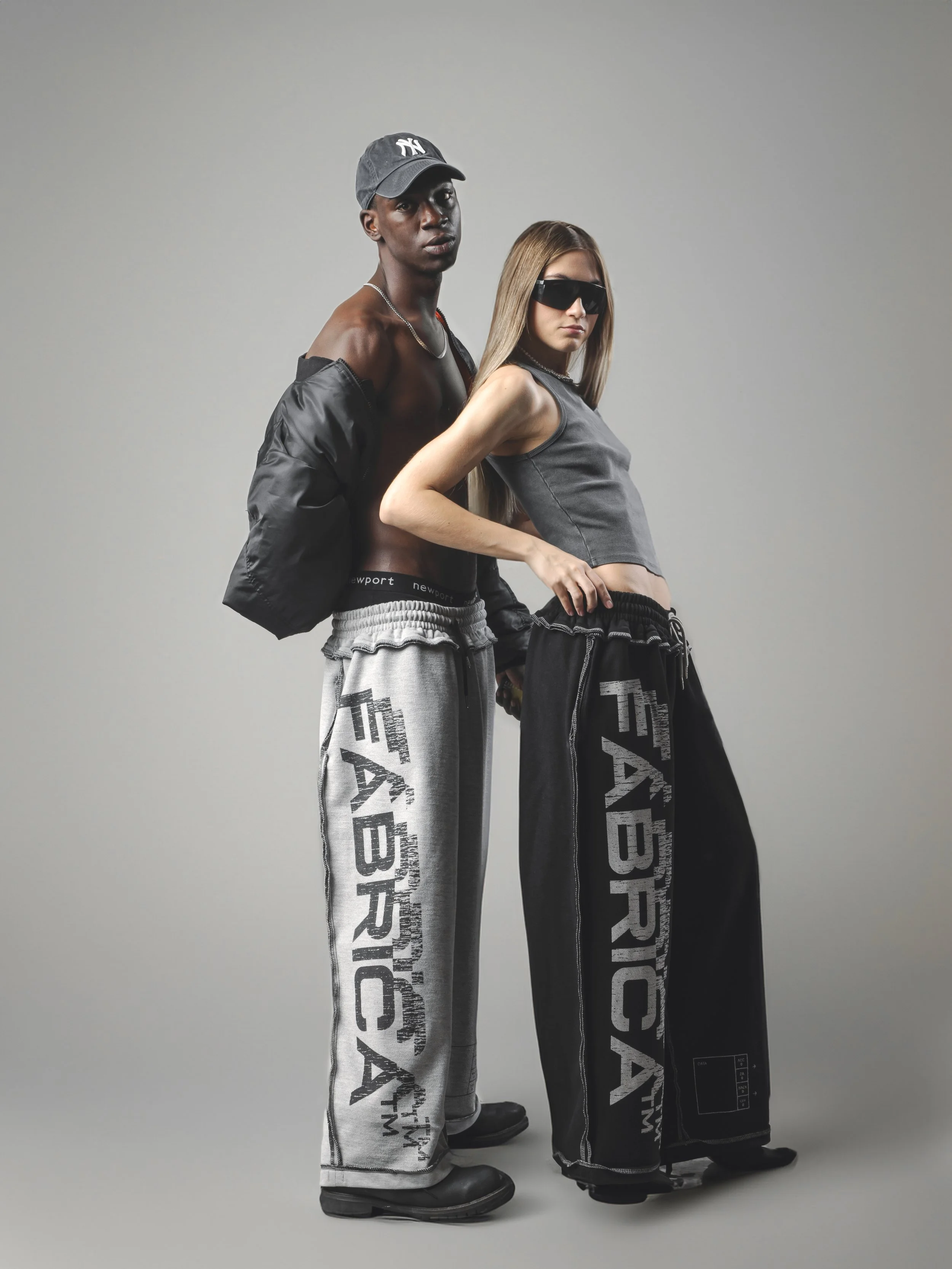 Two models, one male and one female, stand against a plain gray background. The male has dark skin, is shirtless, wearing a baseball cap, oversized pants with text, and black chunky shoes. The female has light skin, long straight hair, wears large bl