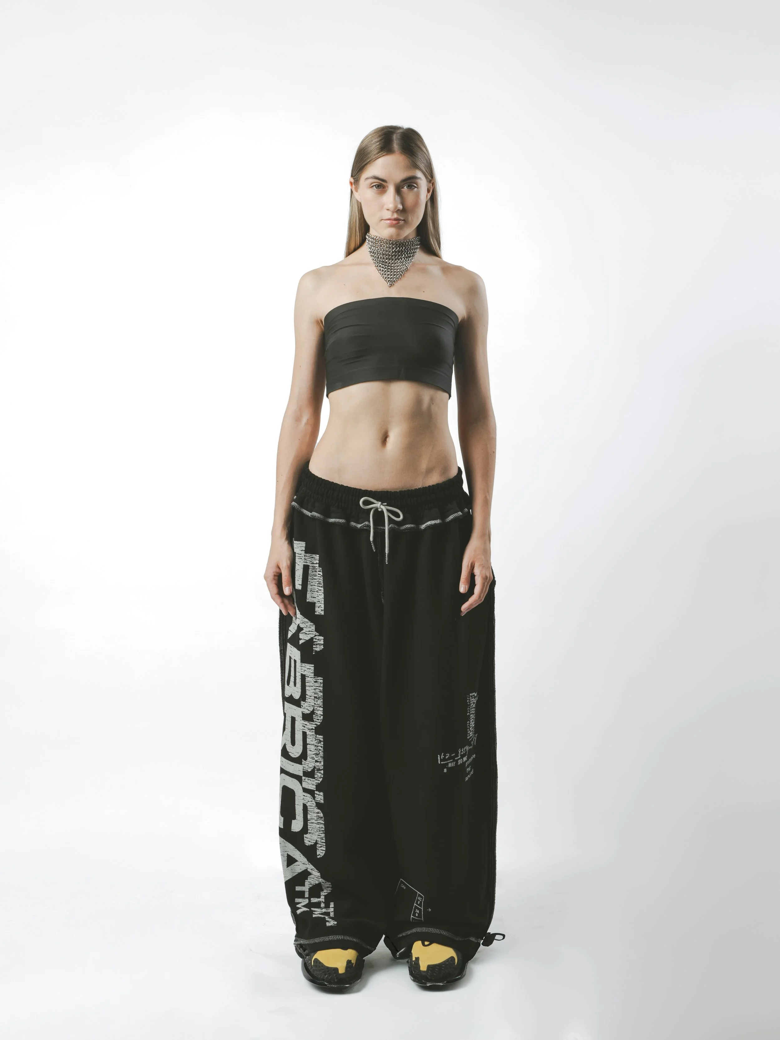 A woman standing against a plain white background, wearing a black strapless tube top, black baggy pants with white text on the side, yellow and black sneakers, and a large, metallic, chainmail necklace.
