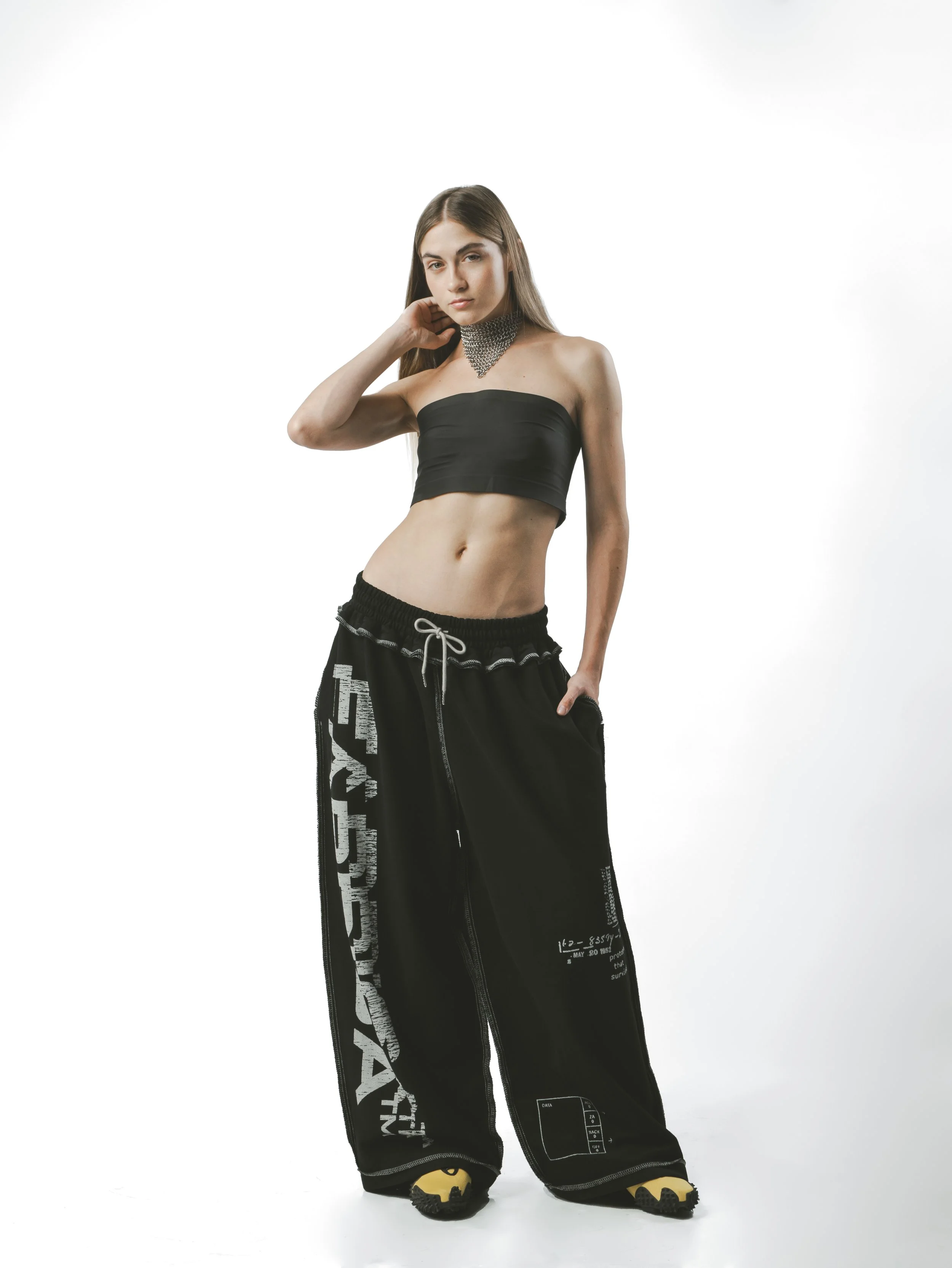 Young woman standing in a white studio wearing a black strapless crop top, black wide-leg pants with white graphics, and yellow shoes with black soles, posing with her left hand on her hip and her right hand touching her neck, gazing into the camera.