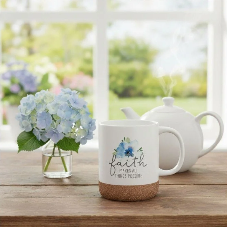 A white mug with a blue flower design and the word 'faith' along with the phrase 'makes all things possible' on a wooden table, accompanied by a white teapot emitting steam and a small glass vase with blue and green hydrangea flowers, with a large window and greenery outside in the background.