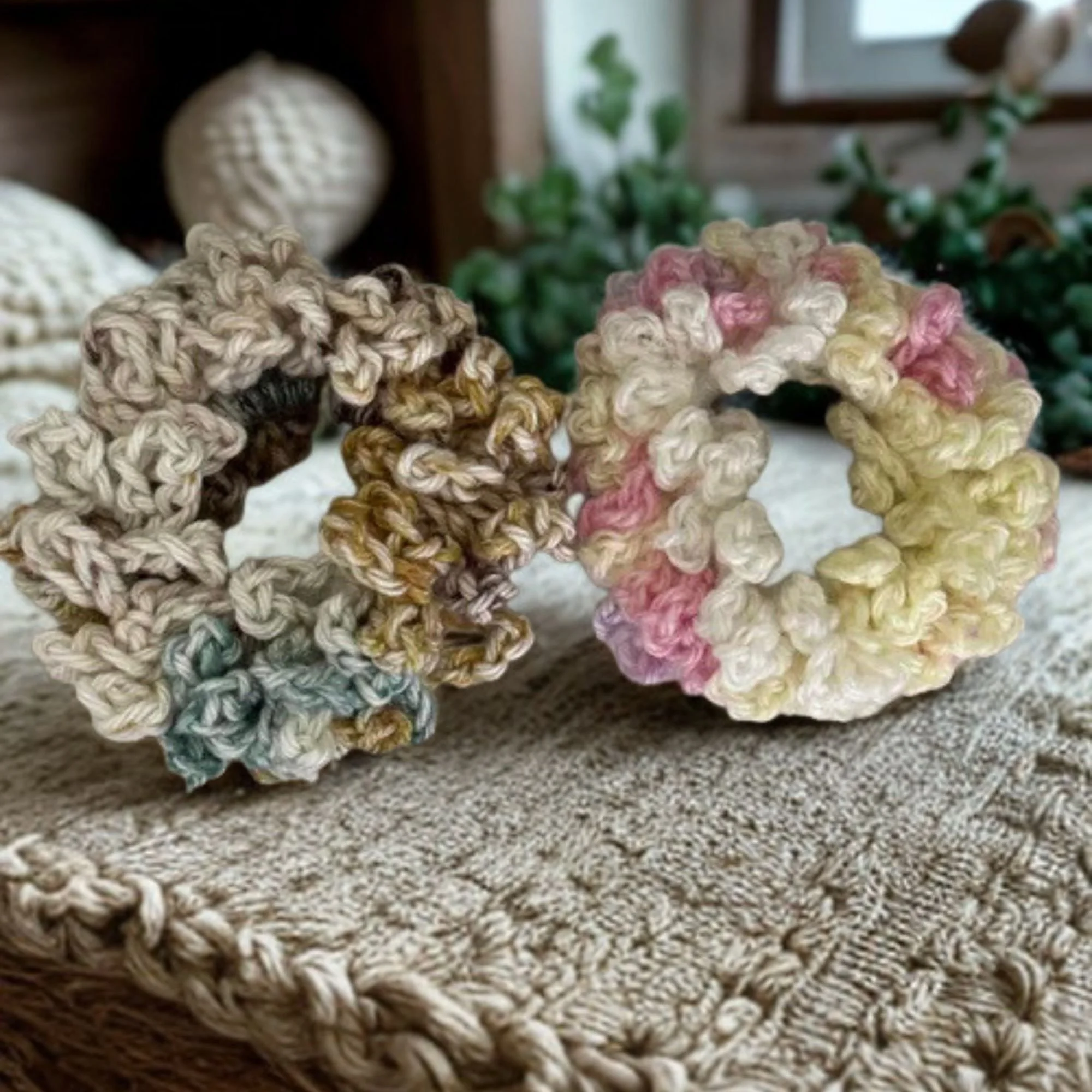 Close-up of woven yarn or crochet pieces in beige, pink, and multicolor, placed on a textured surface with blurred background of plants and a window.