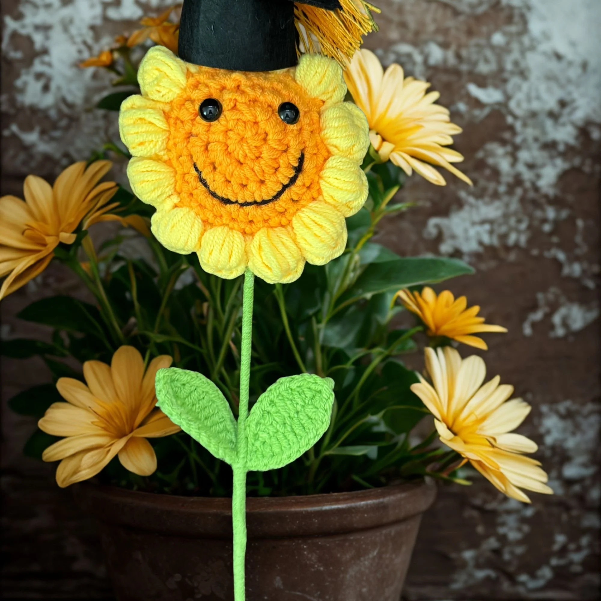 A yellow plush sunflower with a smiling face, black shiny eyes, and an embroidered smile, positioned among real yellow flowers and green leaves in a flower pot.