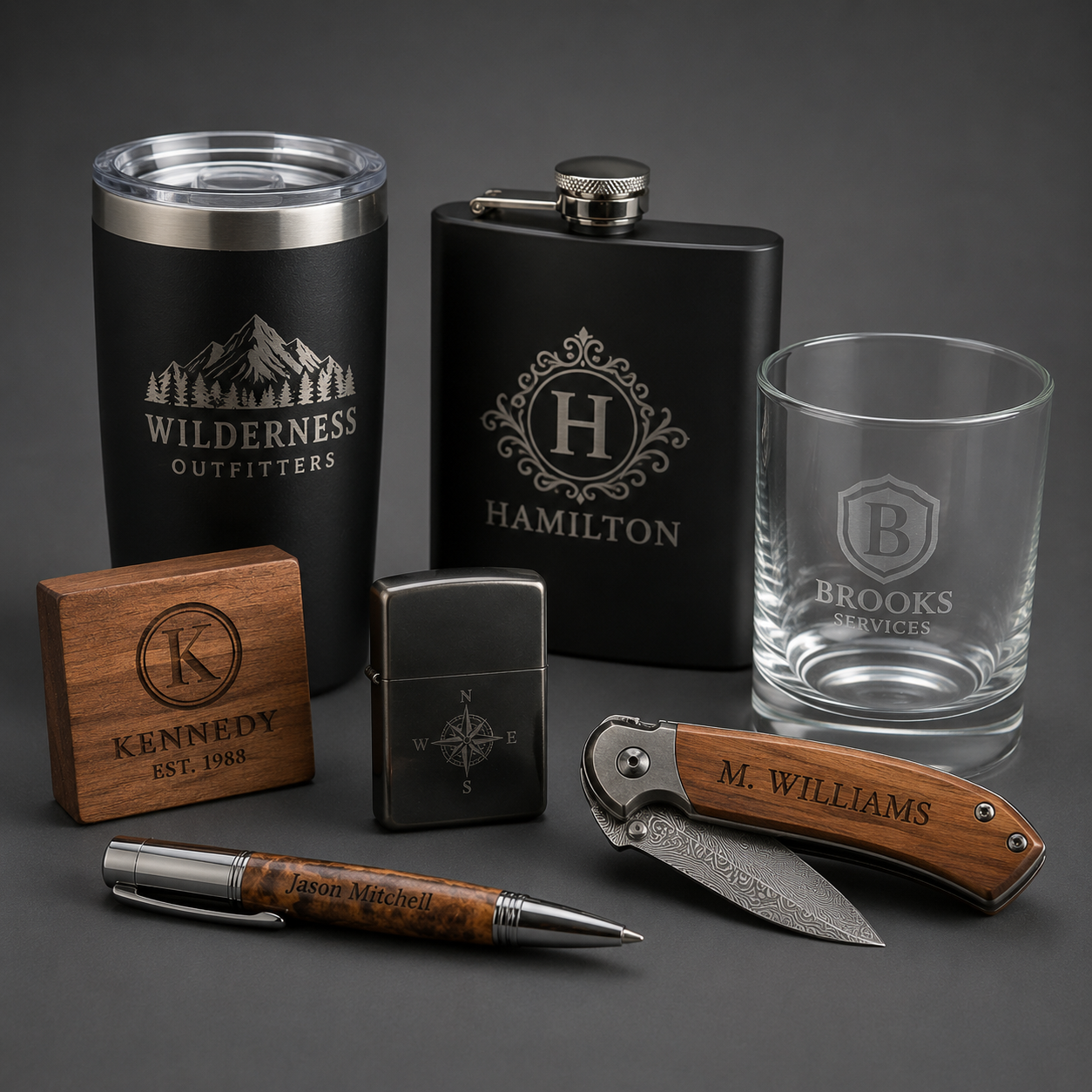 Collection of personalized accessories including a black travel tumbler with wilderness outfitters logo, a black flask with Hamilton logo, a clear glass with Brooks Services logo, a wooden block with Kennedy and established date, a metal compass lighter, a folding pocket knife with engravings, a wooden engraved pen with a name, and a glass in the background.
