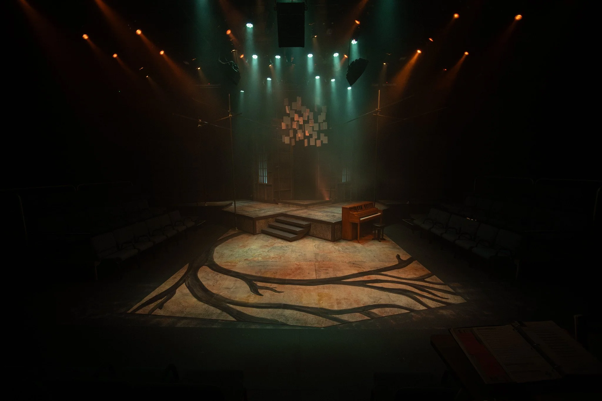Empty theater stage with wooden floor, a small set of stairs, and a piano on the right side. Overhead stage lights cast a colorful glow, and the backdrop features an abstract hanging art installation of white cubes.