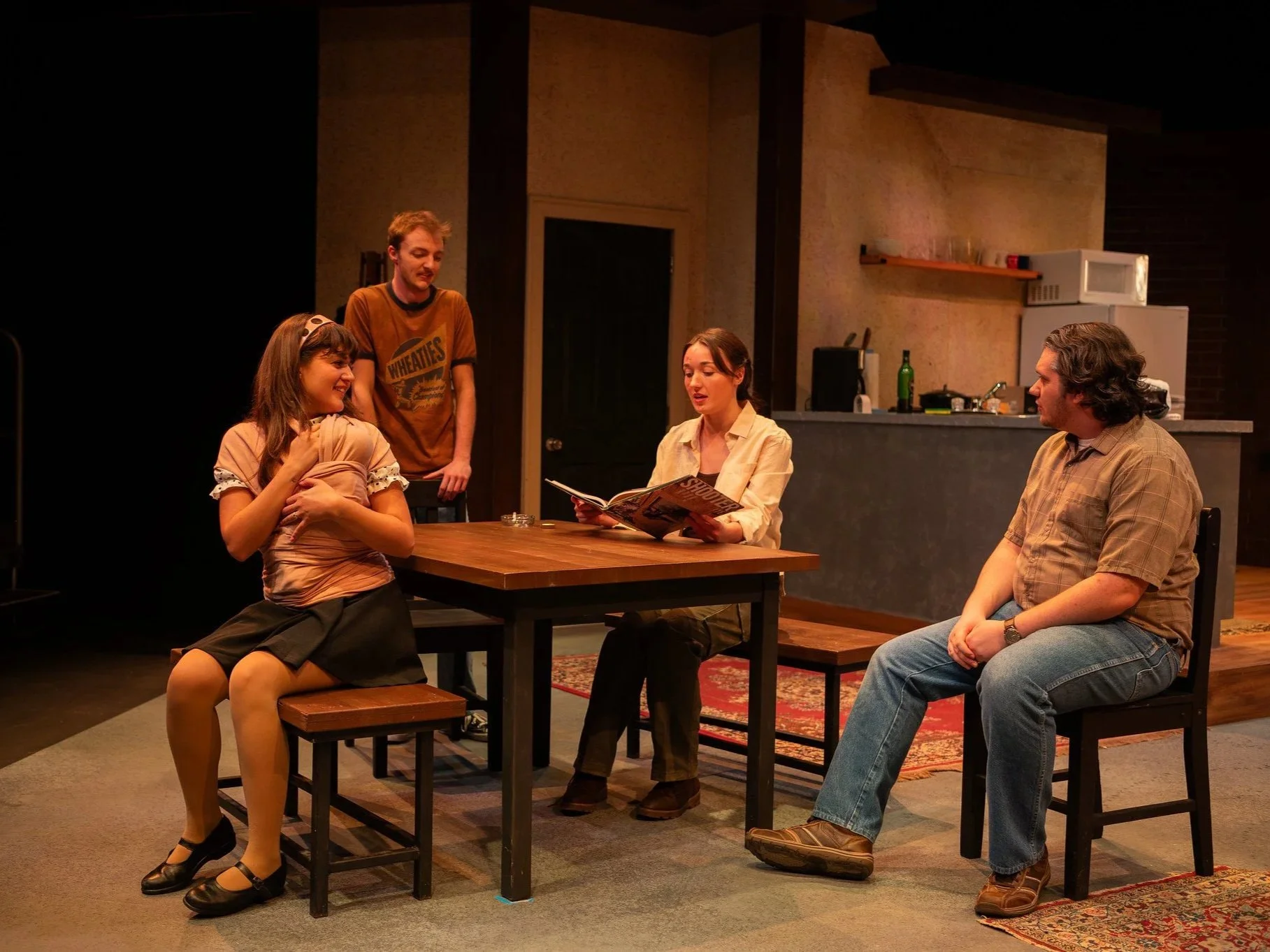 A group of five people engaging in a drama scene or rehearsal in a room resembling a living space or studio. One woman sits on a stool, holding her shirt across her chest, smiling. Another woman sits at the table reading a magazine, while a man sits on a chair to her right, listening. Two men stand behind, observing or participating in the scene, one holding a book or script.