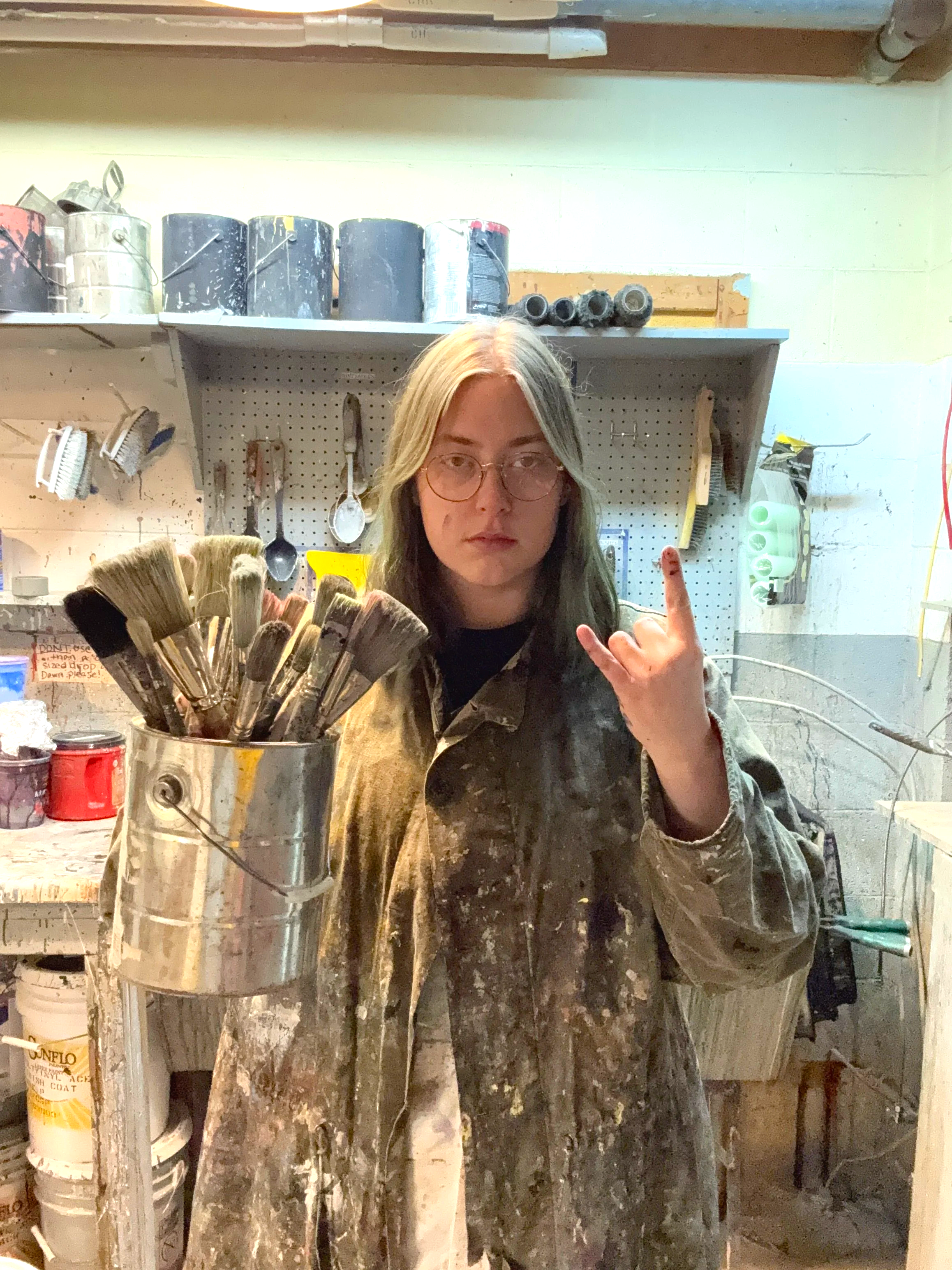 Person with long blonde hair and glasses wearing paint-splattered overalls in an art studio, holding a bucket of paintbrushes, with tools and paint supplies in the background.