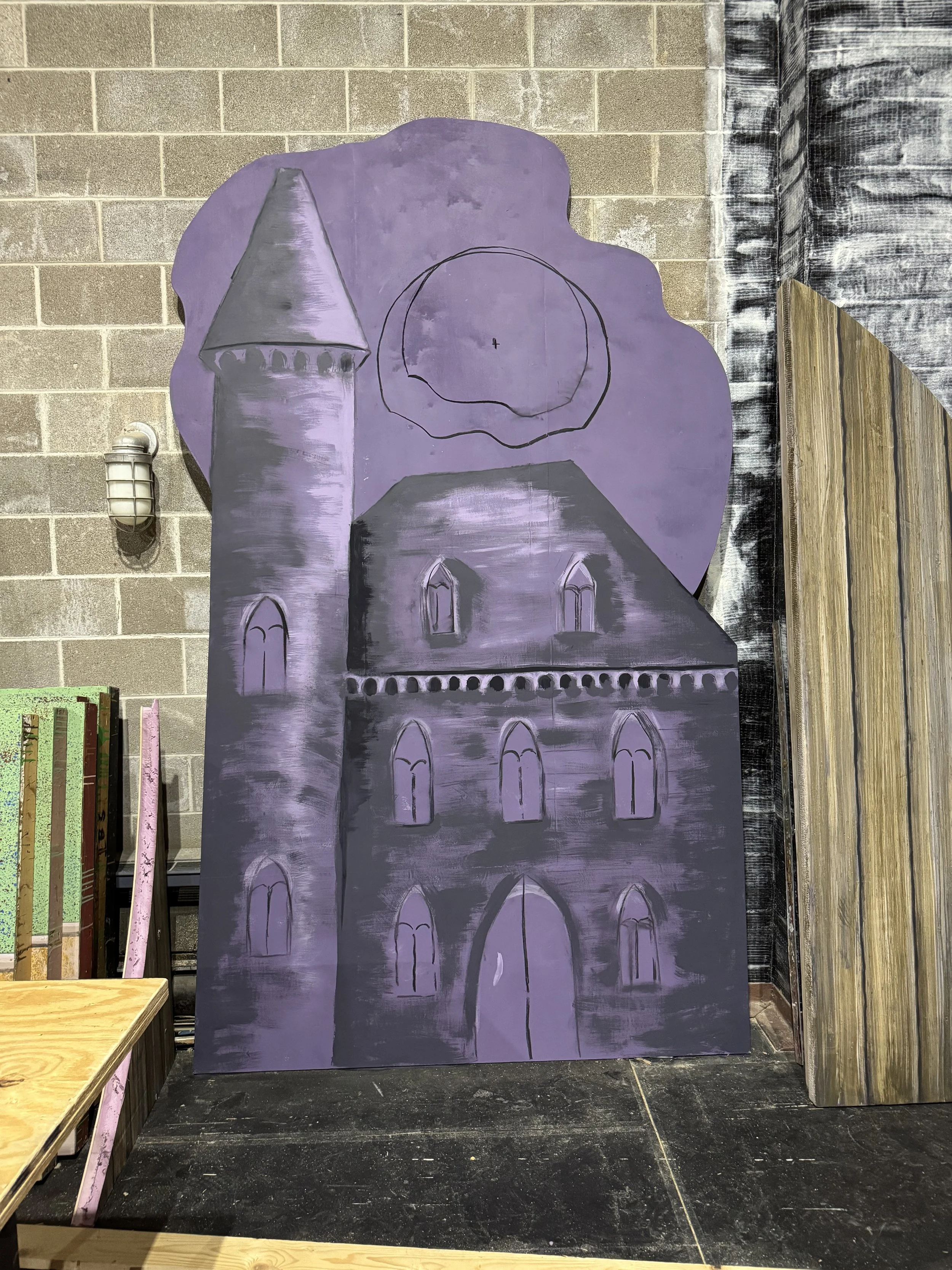 A large purple painted cutout of a castle with towers, windows, and a moon, leaning against a brick wall in an indoor setting.