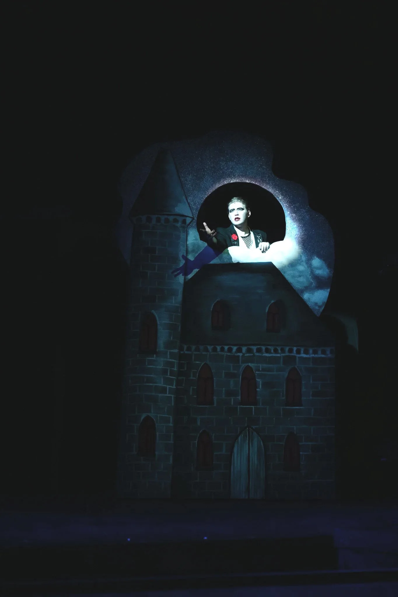 A stage with a castle backdrop, featuring a woman with short hair, makeup, and a black outfit with a red flower, standing inside a tower window.