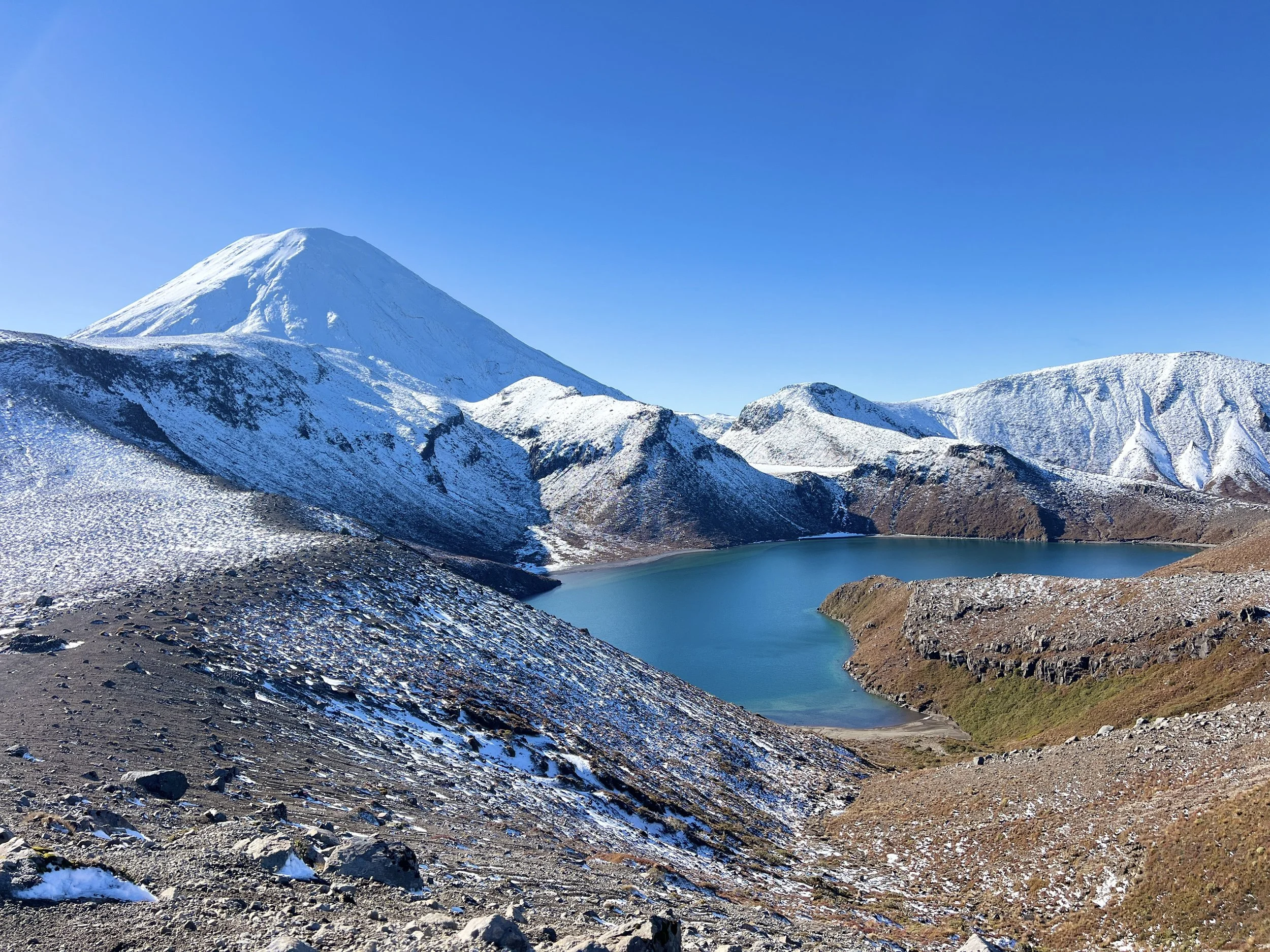 Snow-capped mountain with a blue sky, rocky terrain with patches of snow in foreground, and a turquoise lake at the base of the mountain.
