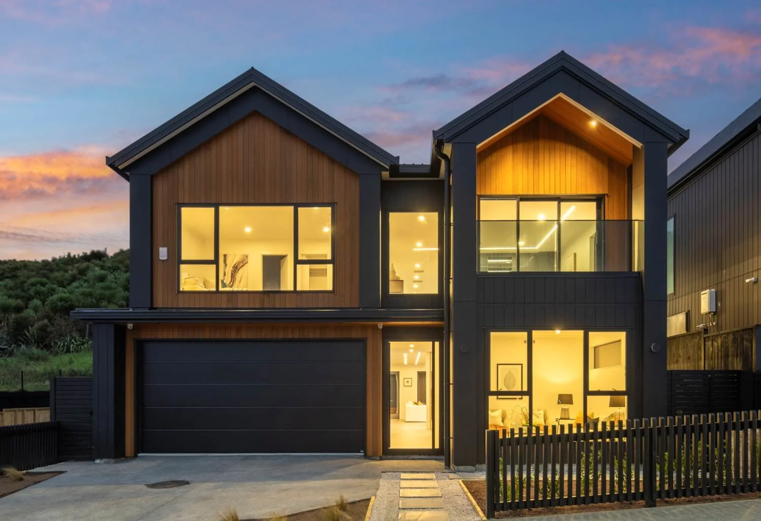 Modern two-story house with large glass windows and warm interior lighting, set against a colorful sunset sky.
