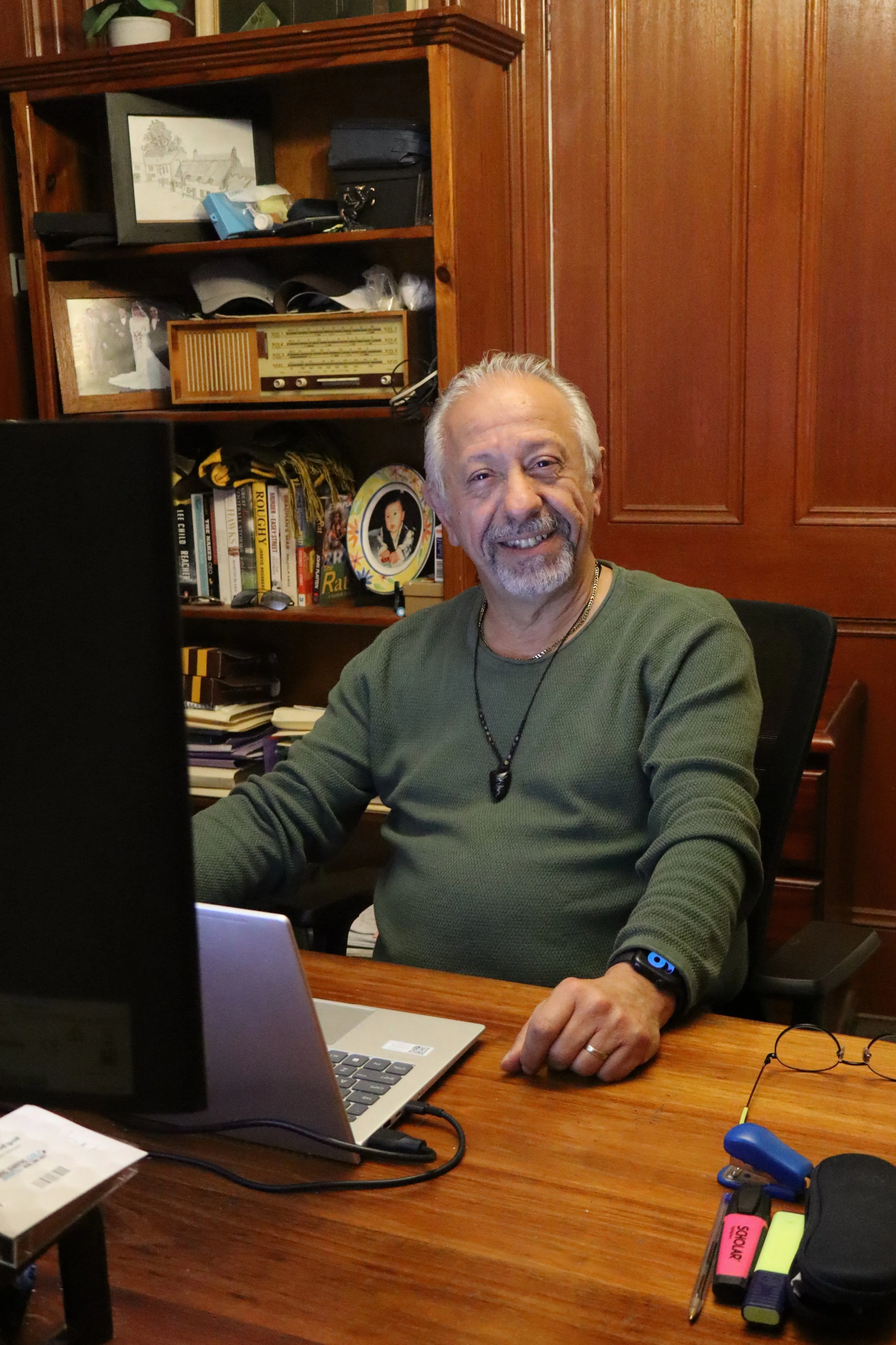 An elderly man with a beard and gray hair smiling and sitting at a wooden desk with a laptop, glasses, and pens, in a room with wooden paneling and bookshelves.