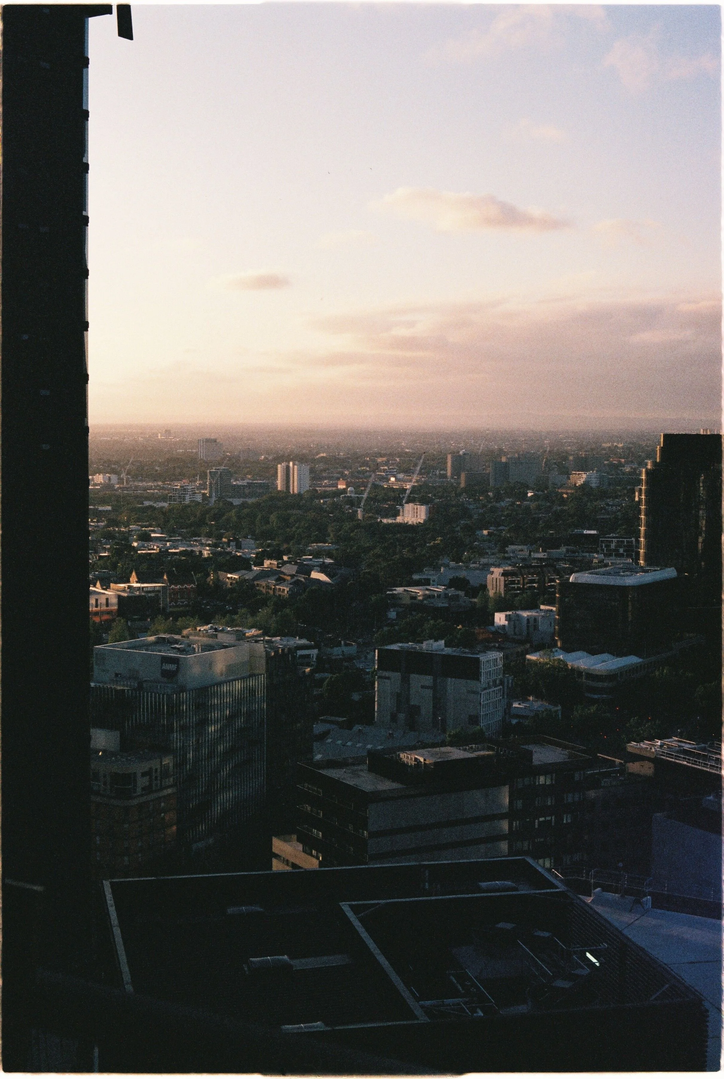 View from a high-rise building showing a cityscape with tall buildings, residential areas, and distant horizon under a partly cloudy sky at sunset.