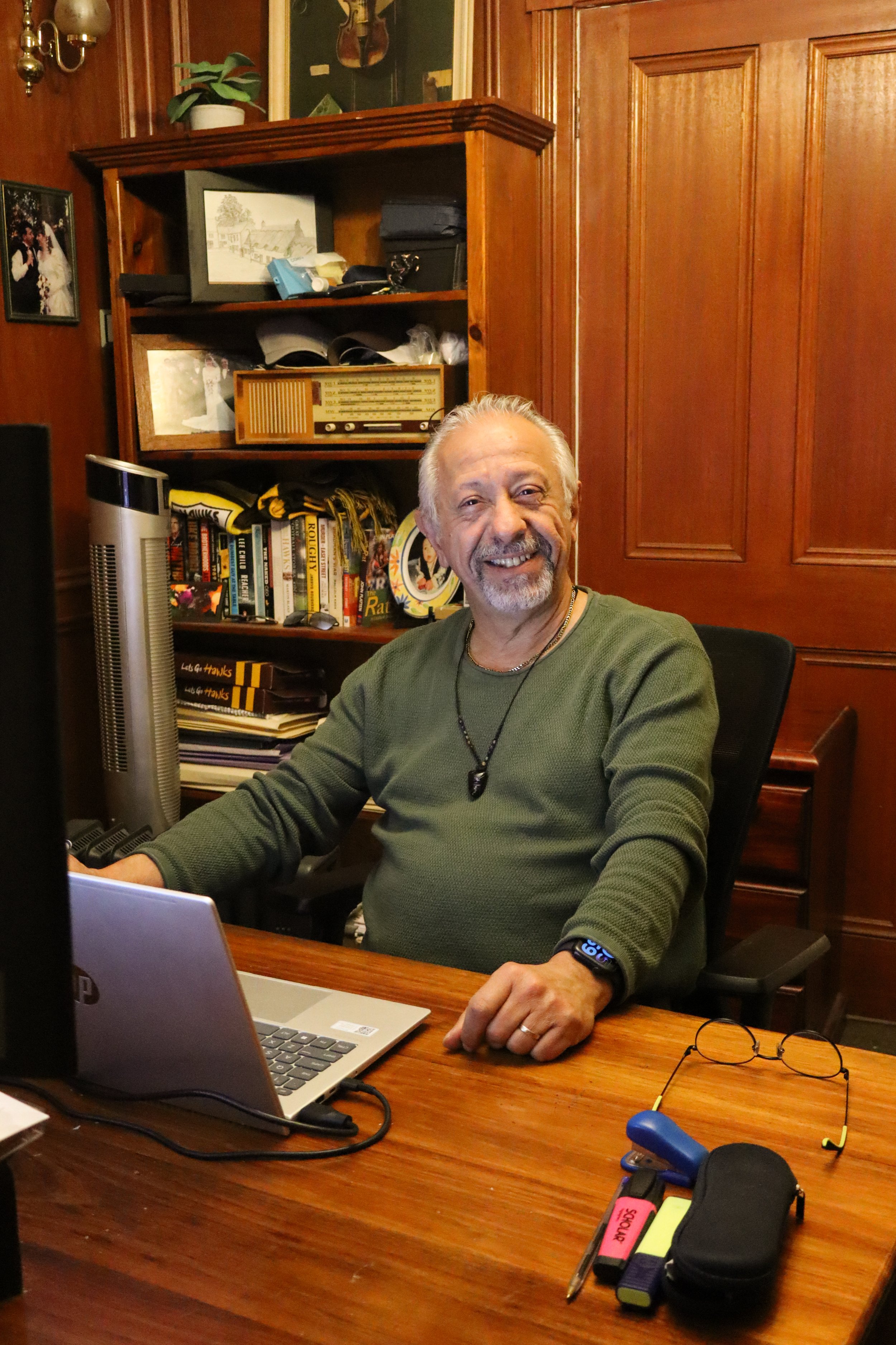 A man with gray hair and a beard smiling at a desk in a wood-paneled room, with a bookshelf, framed photos, and a lamp behind him. There are computer equipment, notebooks, and writing tools on the desk.
