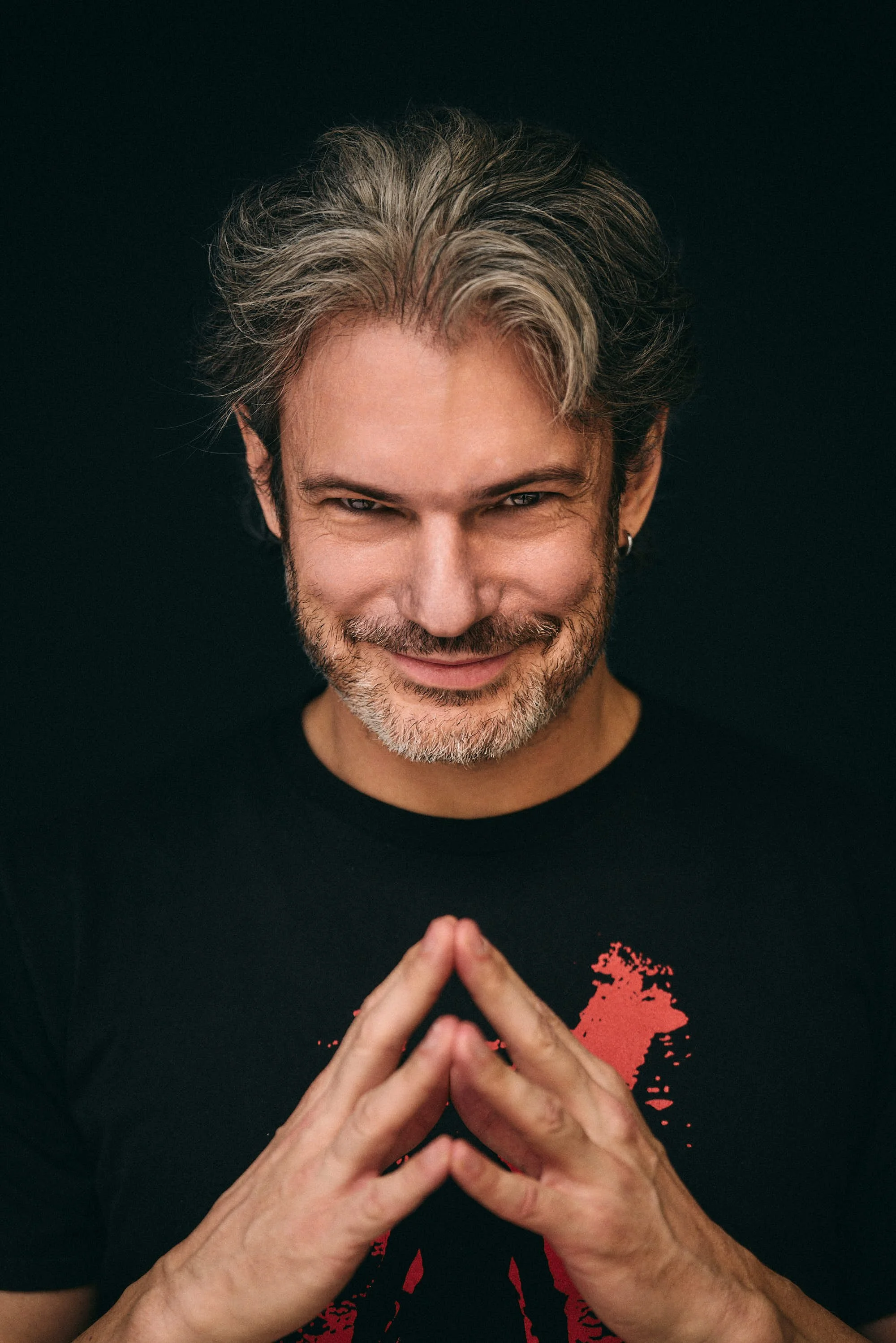 A smiling man with salt-and-pepper hair and beard, wearing a black T-shirt with a red graphic, is making a triangular shape with his fingers in front of him against a black background.