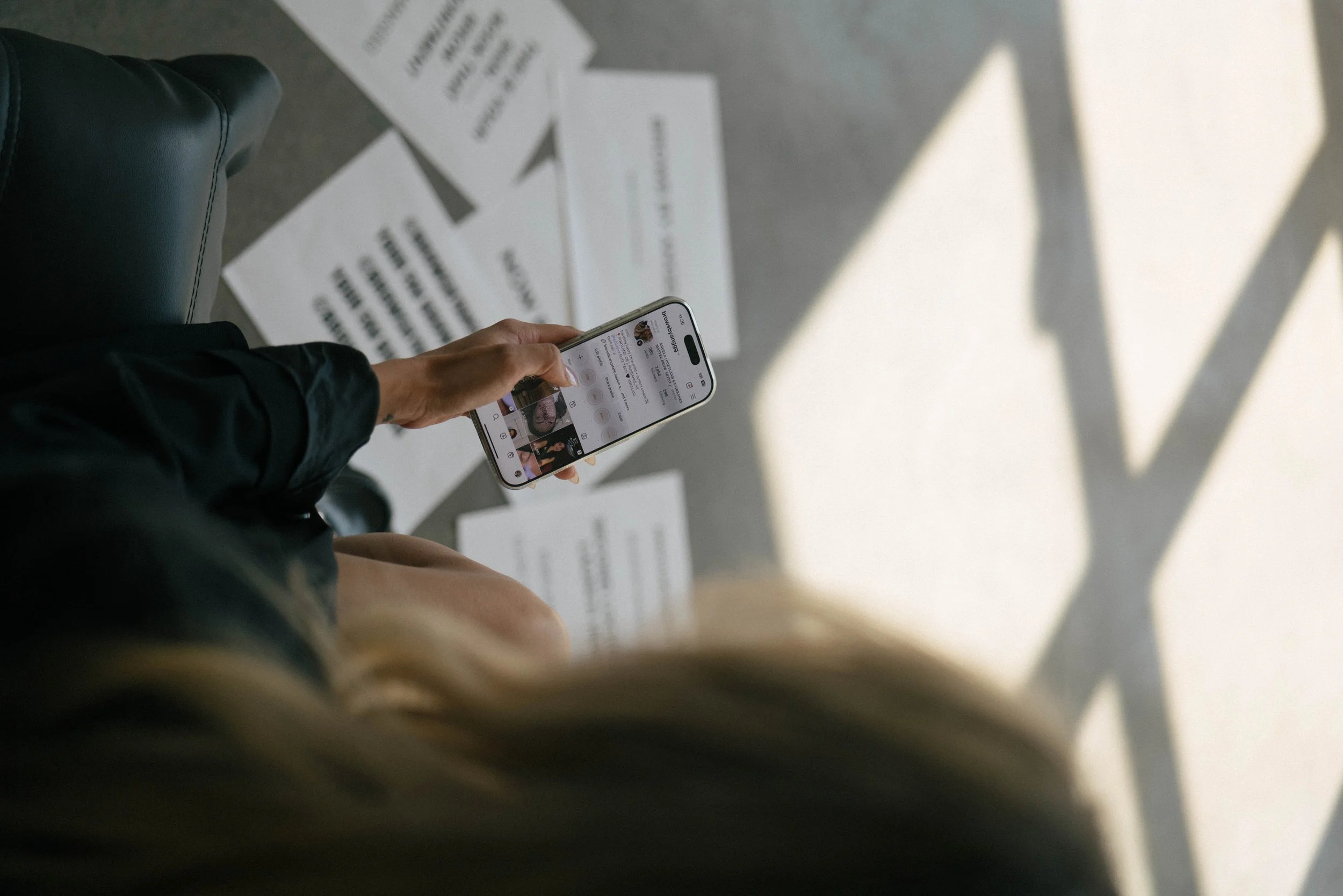 Person holding a smartphone, sitting on a chair with papers scattered on the floor underneath. Shadows cast on the floor.