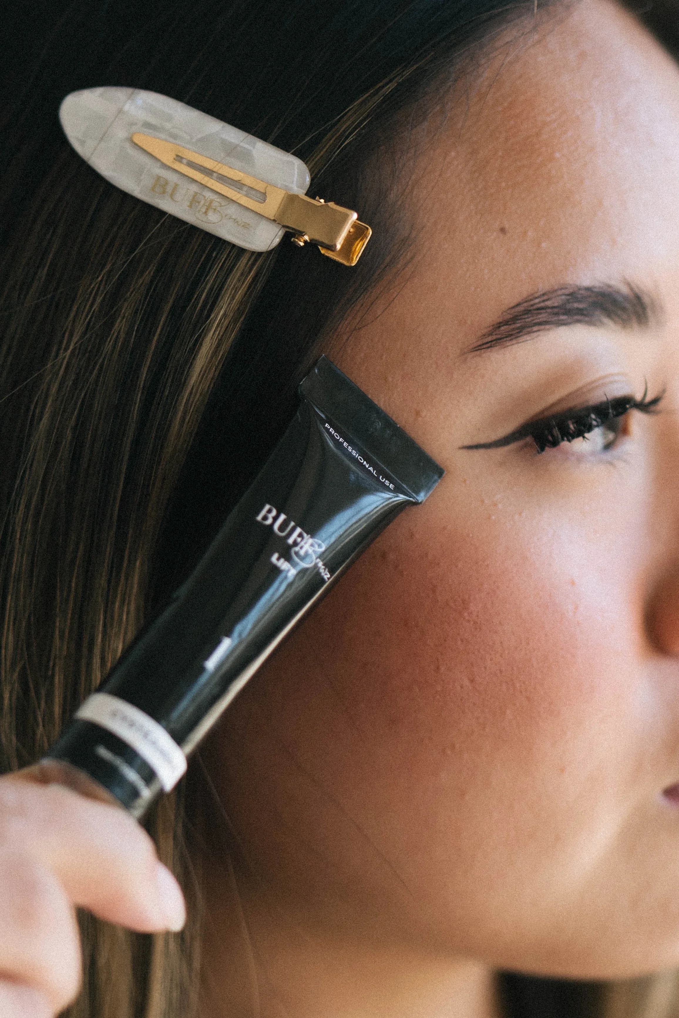 A woman with long brown hair and winged eyeliner holds a tube of Burt's Bees lip balm near her lips, and part of a hair clip is visible in her hair.
