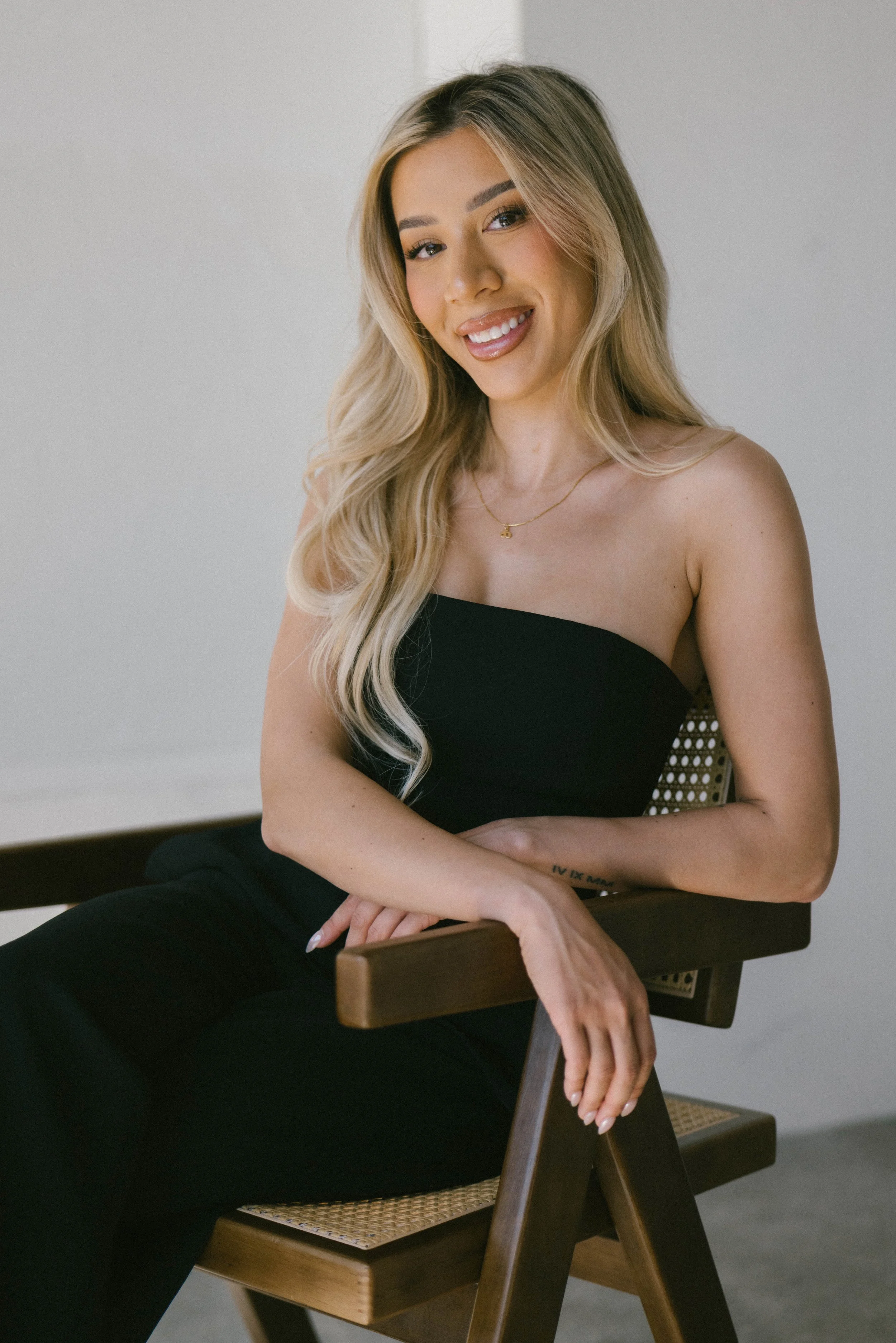 A woman with long blonde hair, wearing a strapless black top, sitting on a wooden chair with a woven seat, smiling and looking at the camera against a plain neutral background.