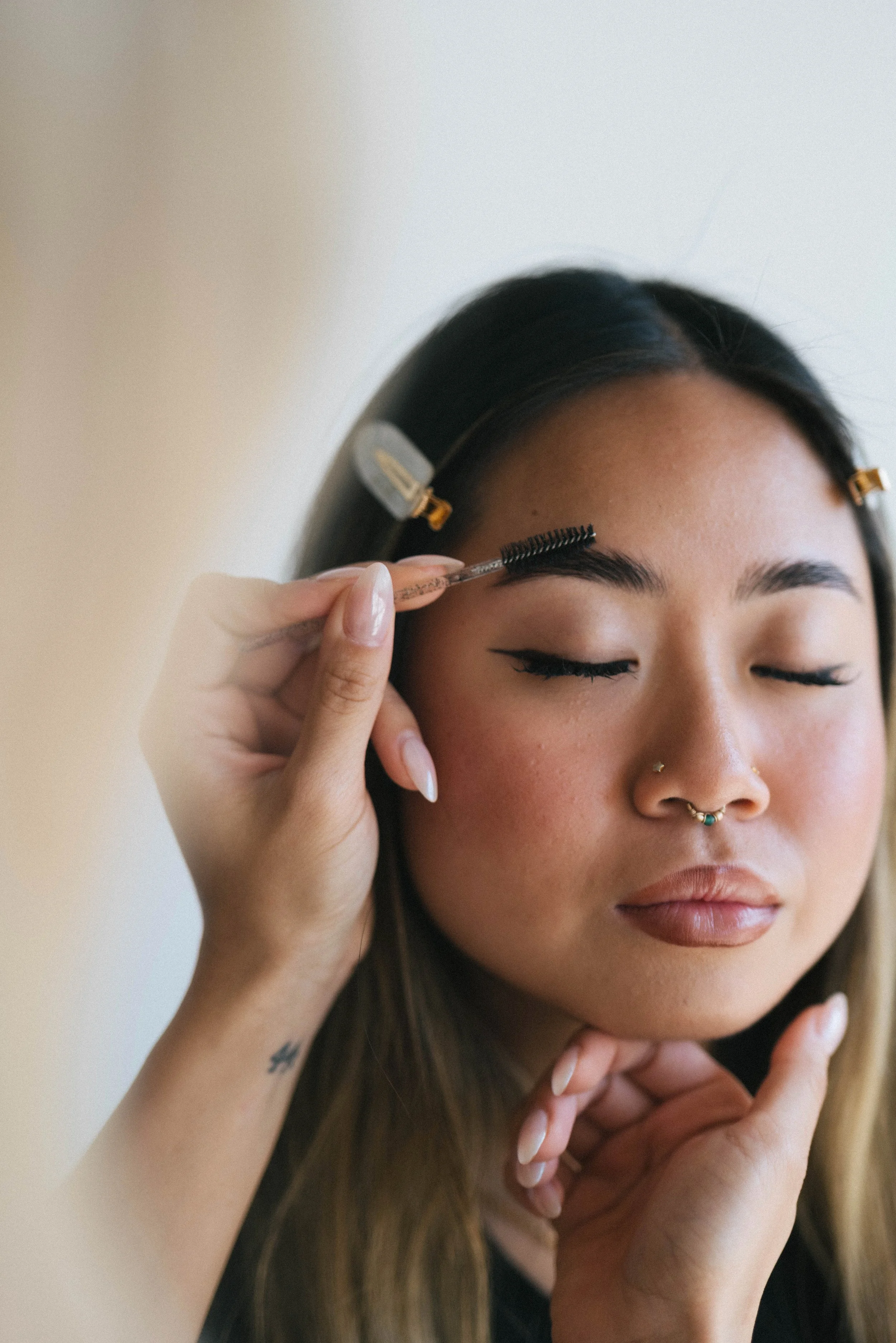 Young woman with long brown hair and septum piercing having her eyebrows groomed using a spoolie brush, with hair clips in her hair and her eyes closed.