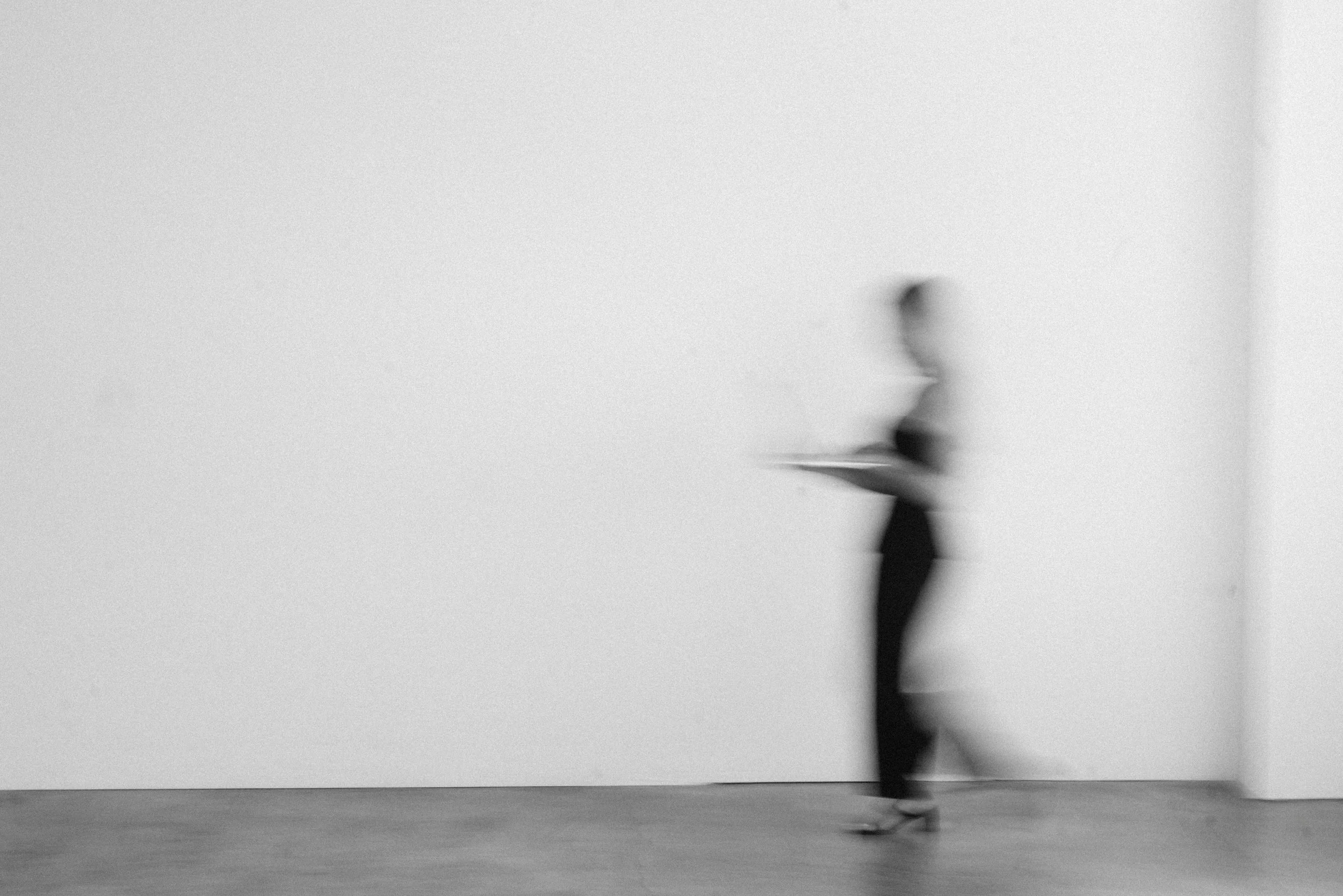 A blurred person walking in a gallery with plain white walls and a light floor.