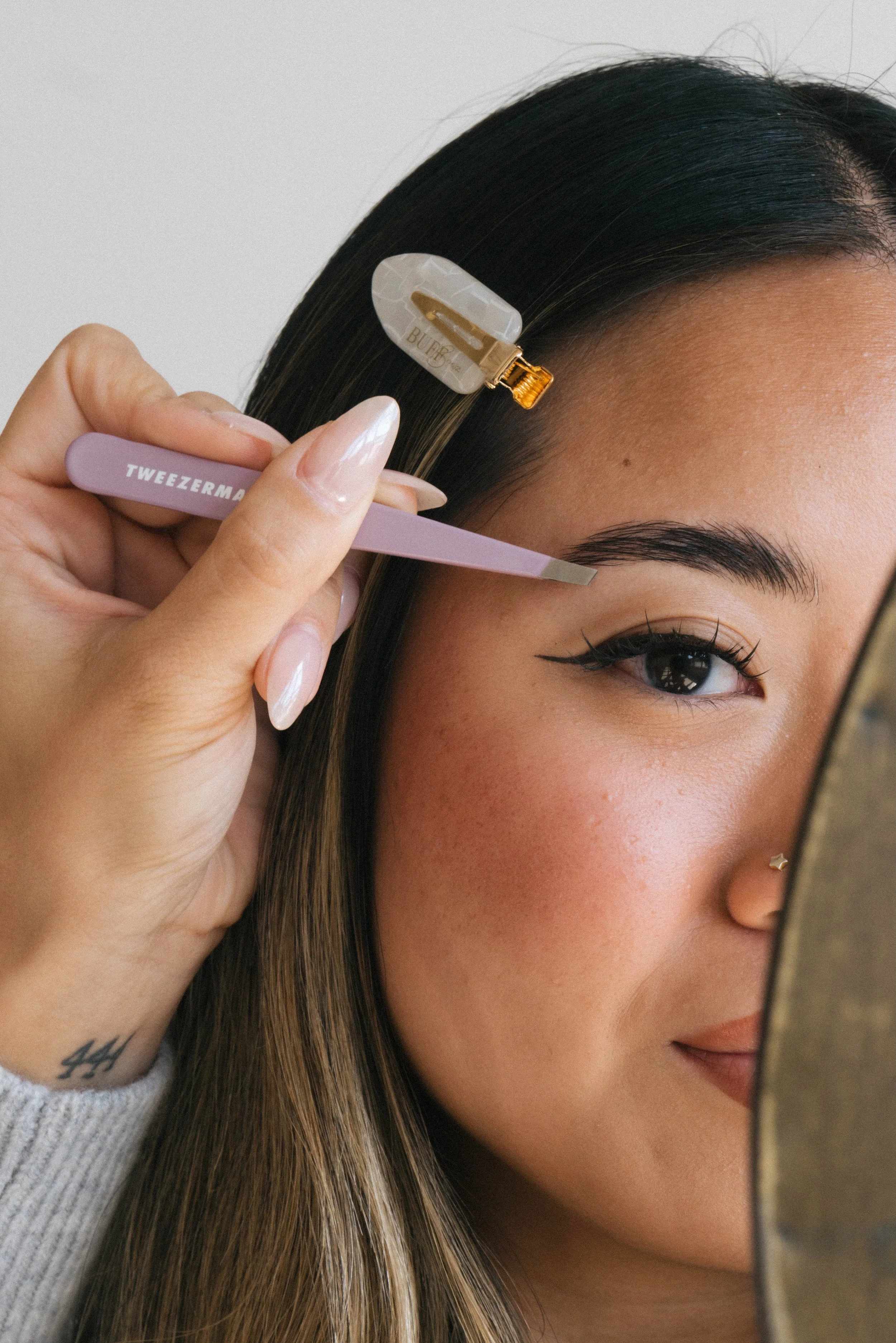 A woman with long, straight hair is using a small metal tool to tweeze her eyebrows while looking into a mirror. She has a gold hair clip holding back part of her hair and a small star-shaped piercing on her earlobe.