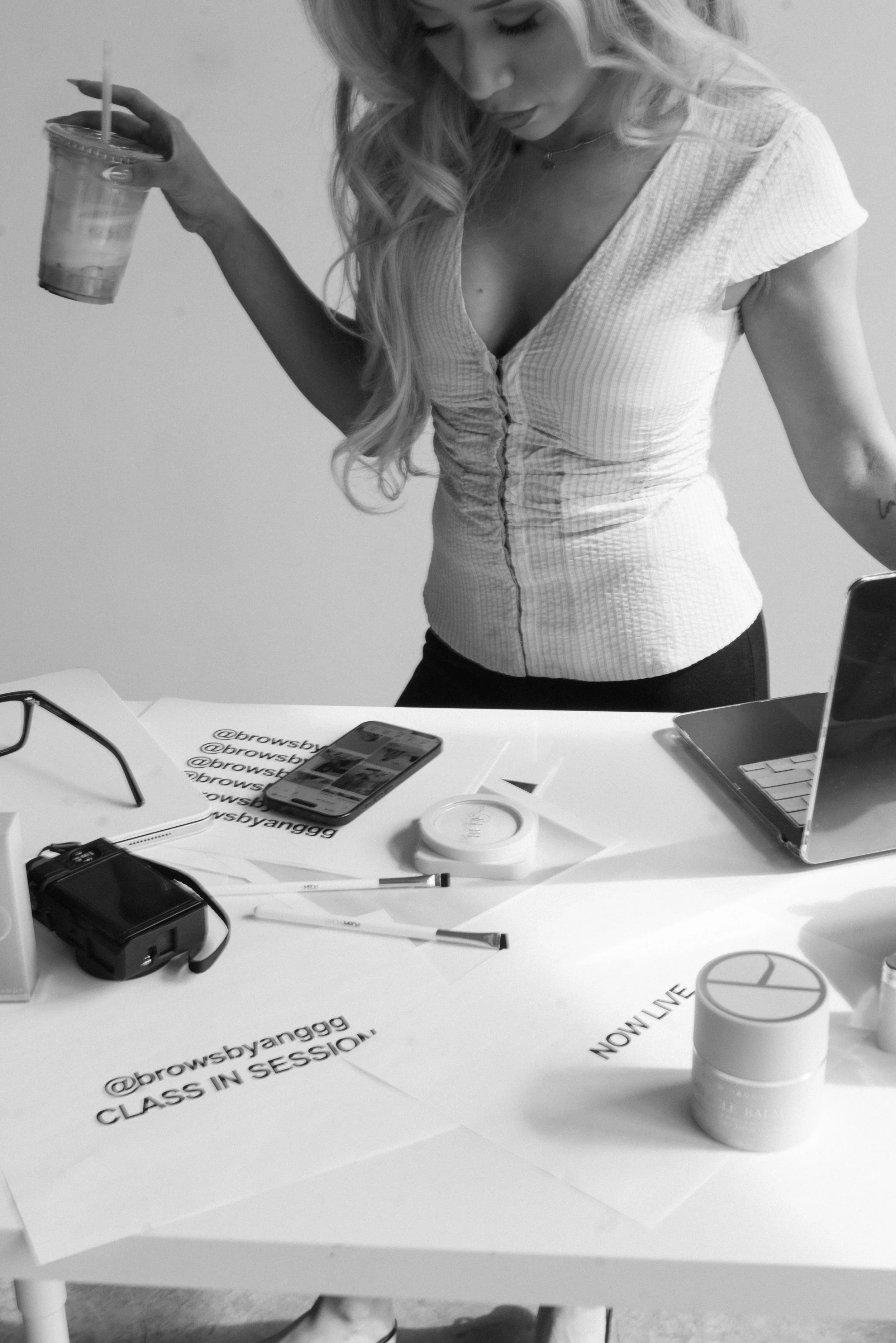 A woman with long wavy hair holding a cold beverage in a clear plastic cup with a straw, wearing a fitted, ruched, short-sleeve top, standing at a cluttered white table with various objects, including a smartphone, papers, a container, a pair of glasses, a pen, and electronic devices.