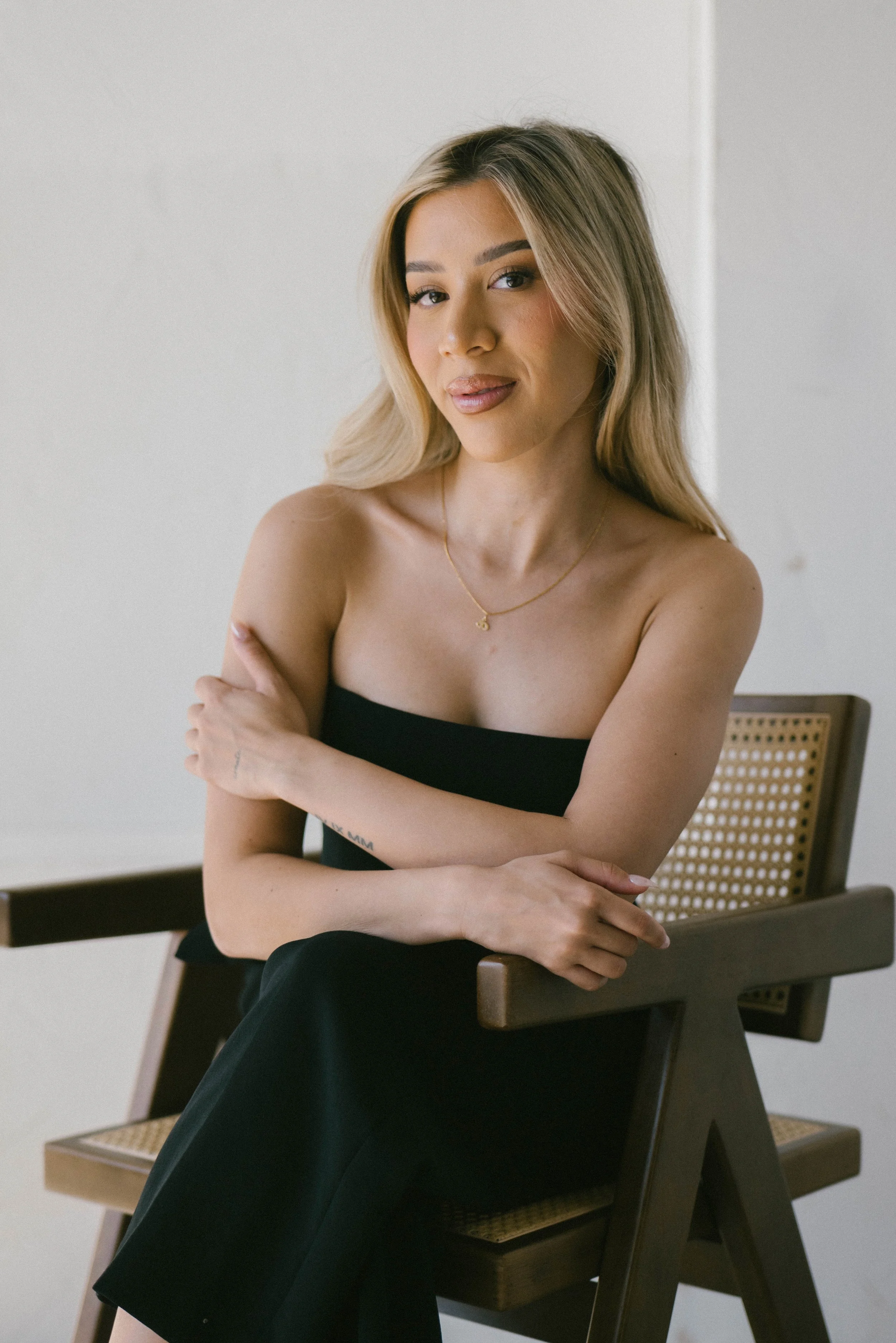 A woman with blonde hair sitting on a wooden chair with a cane back, wearing a black tube top, and a delicate gold necklace, posing with one arm crossed over her chest and the other resting on her lap, in a minimalistic room with light-colored walls.