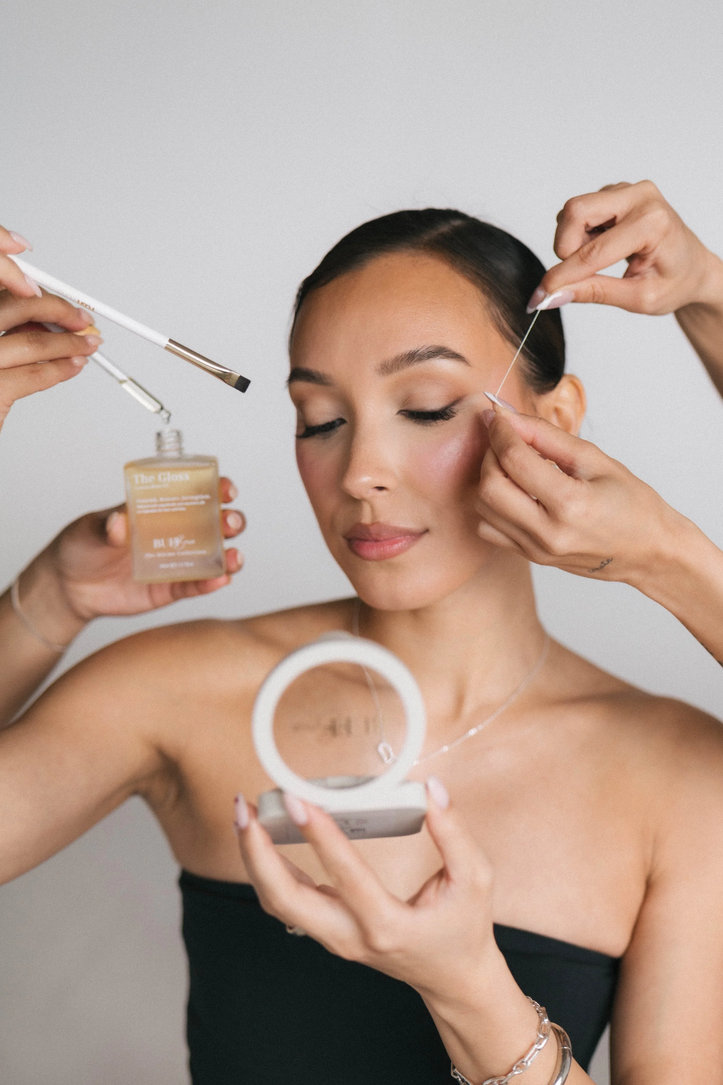 A woman with dark hair getting her eye makeup done by a makeup artist, who is holding a liquid eye liner and brush. The woman is holding a compact mirror, and she is wearing a black top and jewelry. The background is plain and light-colored.