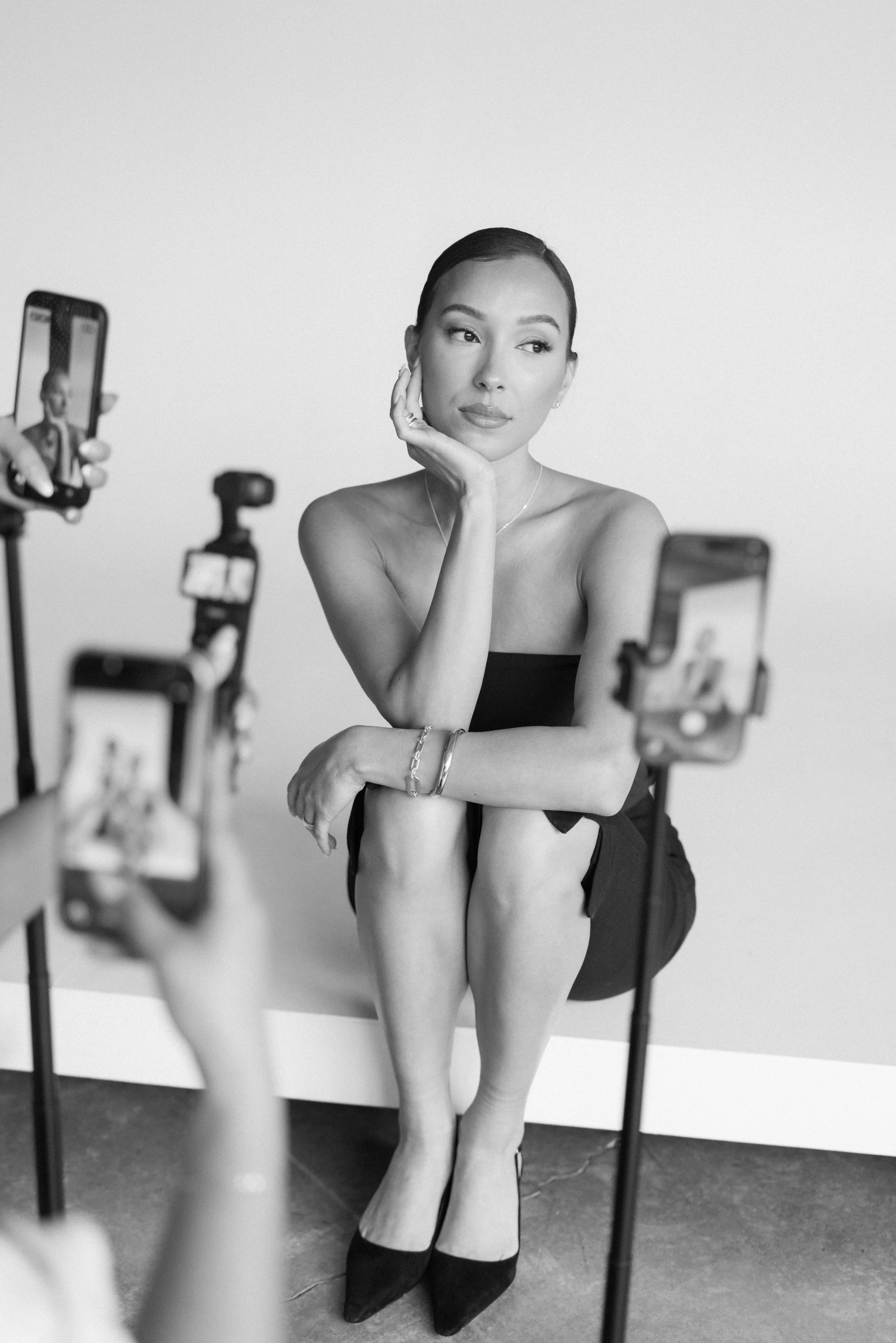 A woman with short hair, wearing a strapless top and high heels, posing in front of a plain white background while multiple smartphones on tripods record her.