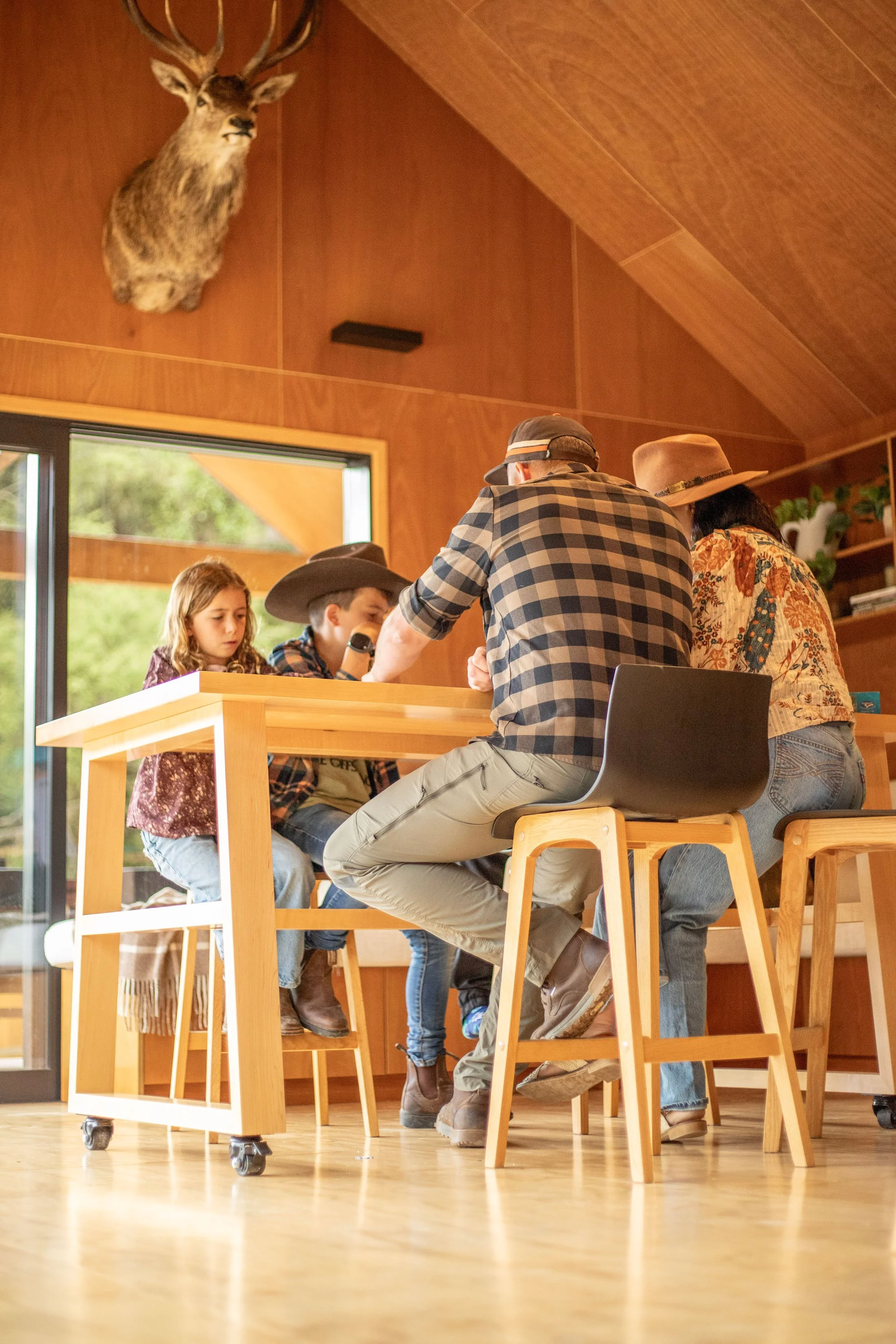 A group of people, including children and adults, gathered around a wooden table, engaging in an activity. A mounted deer head is displayed on the wood-paneled wall above them. Natural light coming through large windows illuminates the room, which ha