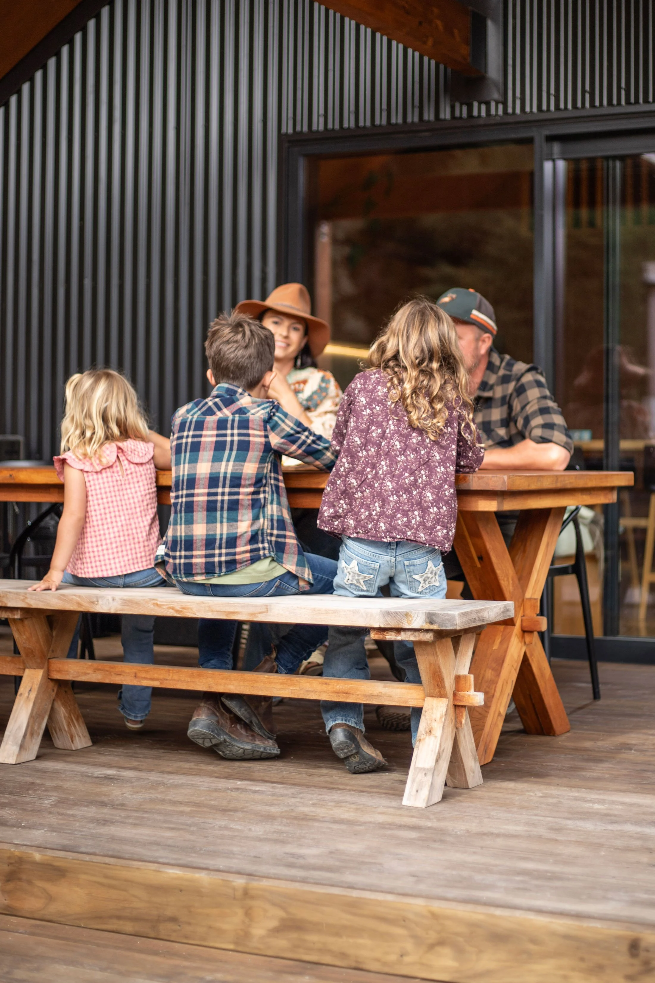Group of children and adults sitting and standing around a wooden picnic table on a porch or deck, engaged in conversation, with a modern-style building with large windows and black vertical siding in the background.