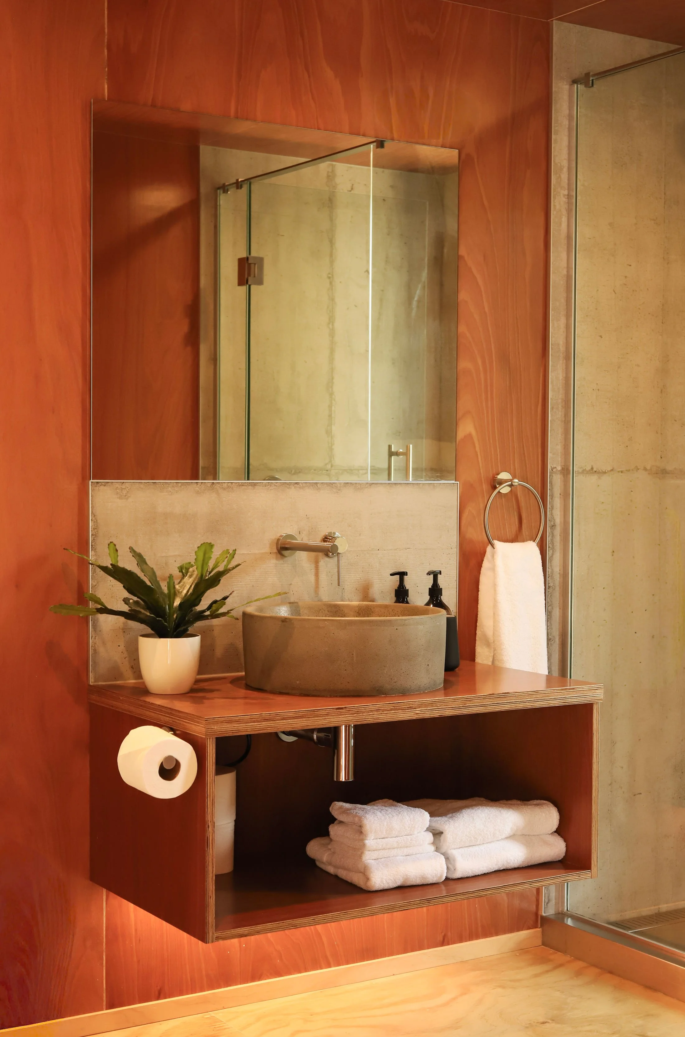 A modern bathroom with a floating wooden vanity, a round concrete sink, a large mirror, a potted plant, a black soap dispenser, a towel ring with a white towel, and stacked white towels on an open shelf. There is a glass-enclosed shower with concrete