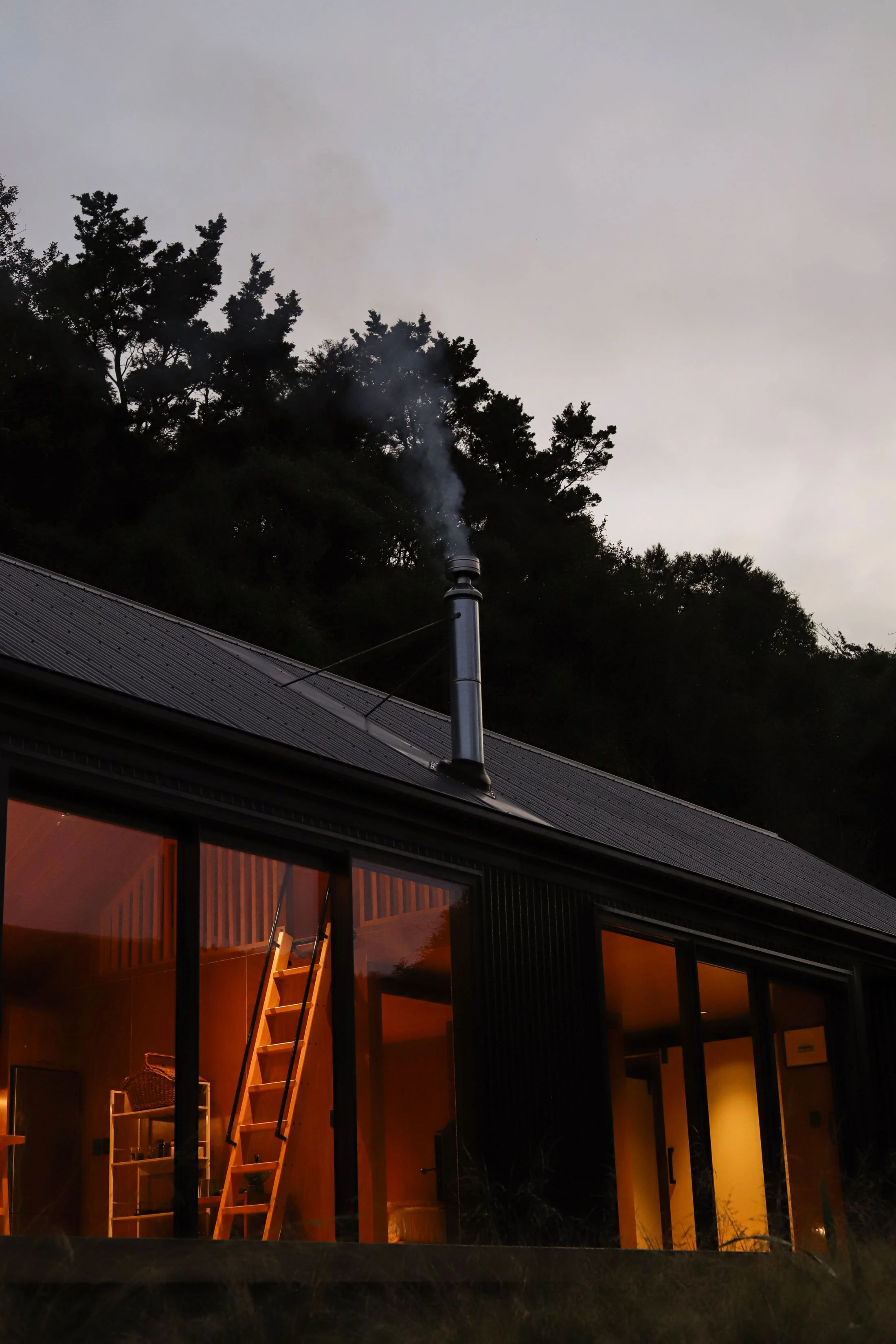 A house with large glass windows and a metal roof, glowing warmly inside, with a staircase leading up to a door inside, and trees and a chimney with smoke outside during dusk.