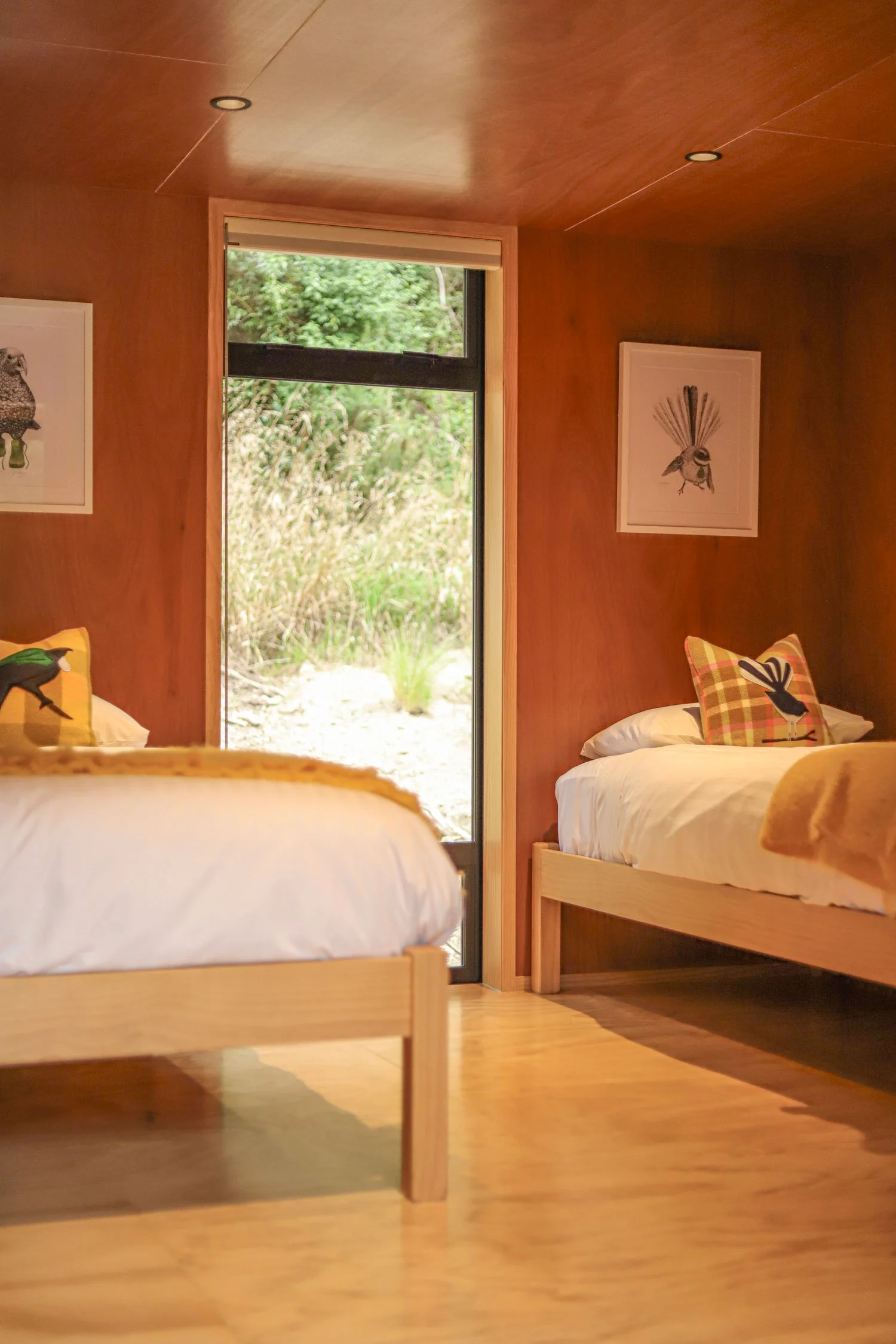 Two wooden beds with white bedding and colorful pillows, inside a cozy room with wooden walls and ceiling, large window showing outdoor greenery.