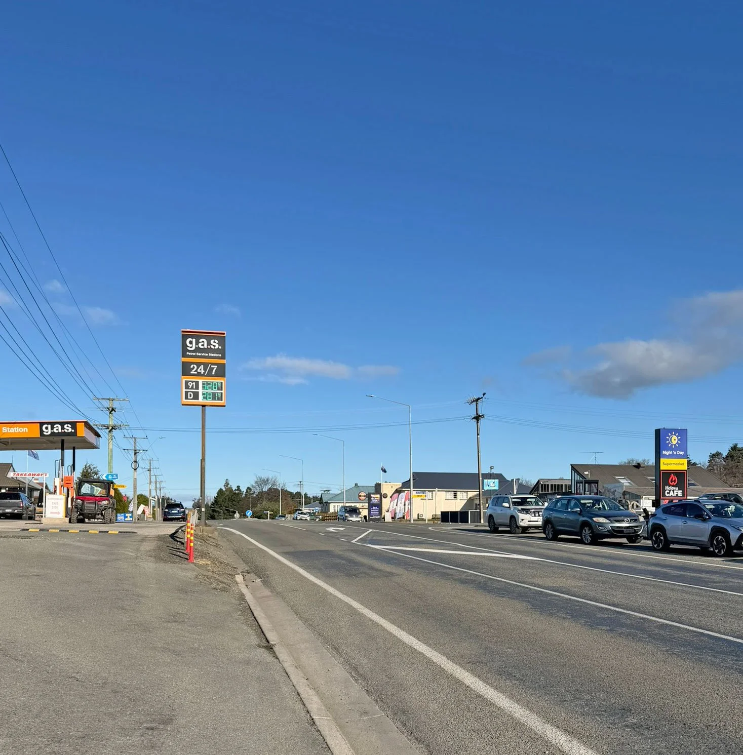 A street view of a small commercial area with a gas station, stores, and parked cars under a clear blue sky. Signs display gas prices and store names.